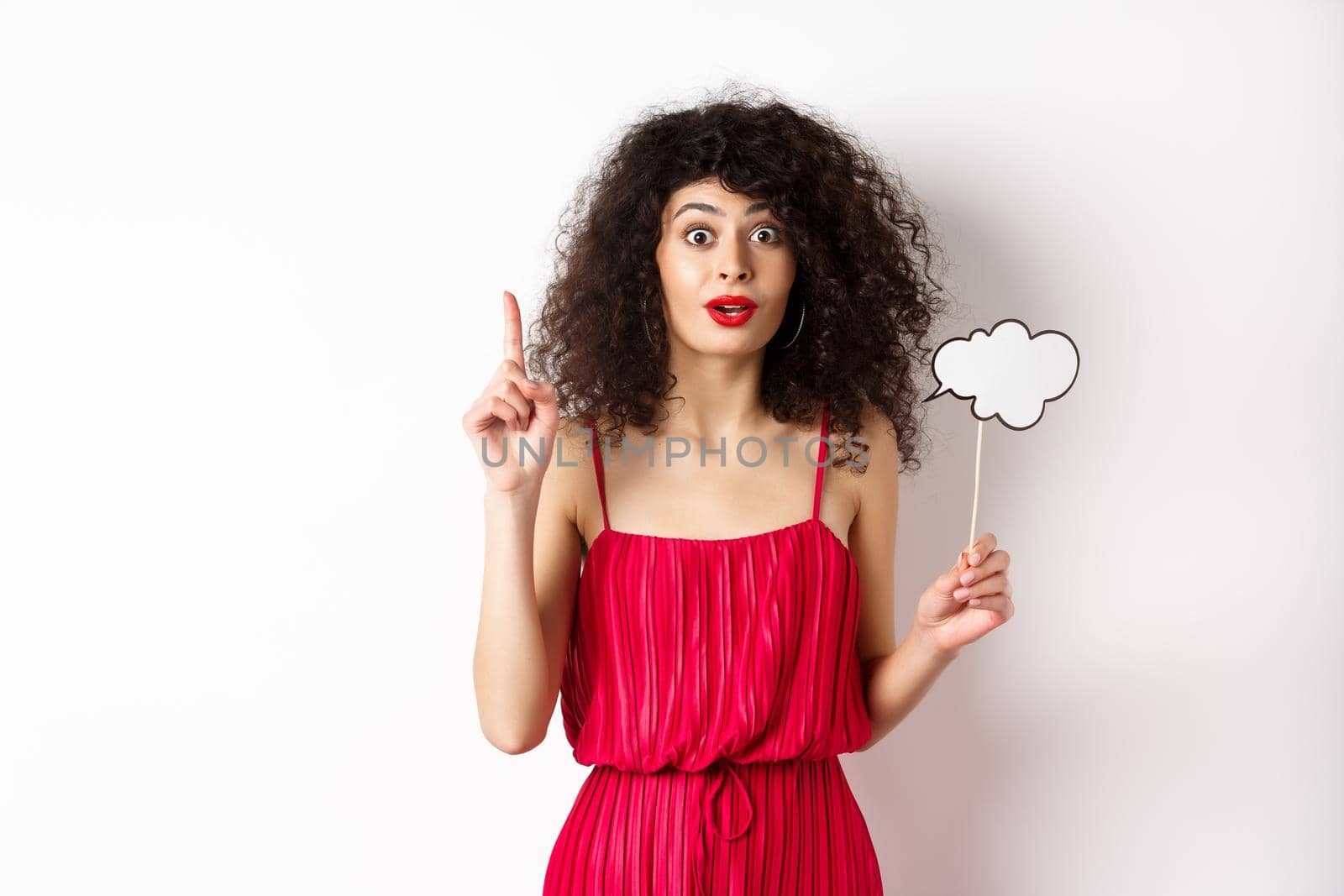 Cute caucasian lady in red dress, holding comment cloud and raising finger, pitching an idea and smiling, suggesting something, standing over white background.