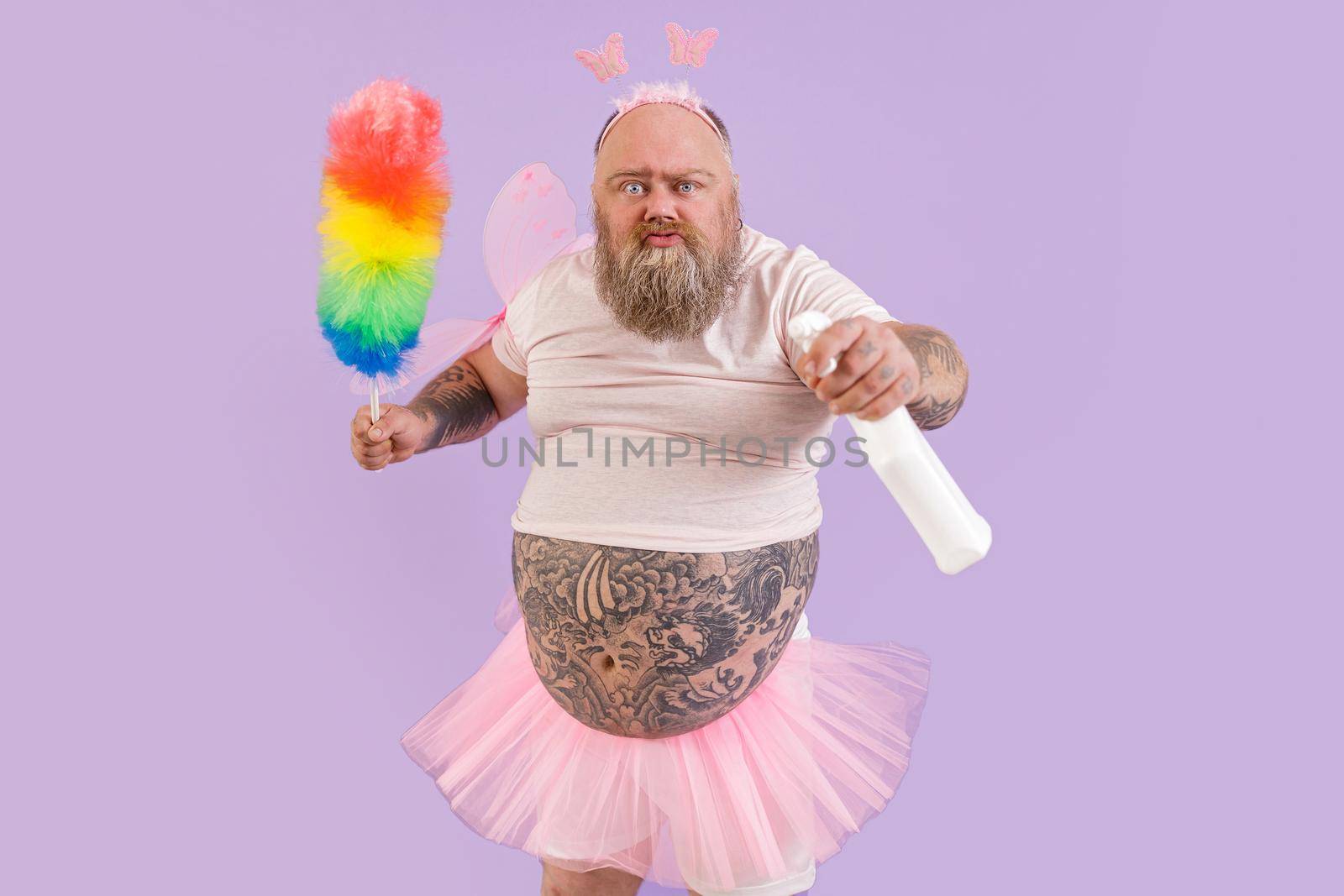 Funny mature male person with overweight in fairy costume with duster and spray bottle poses on purple background in studio