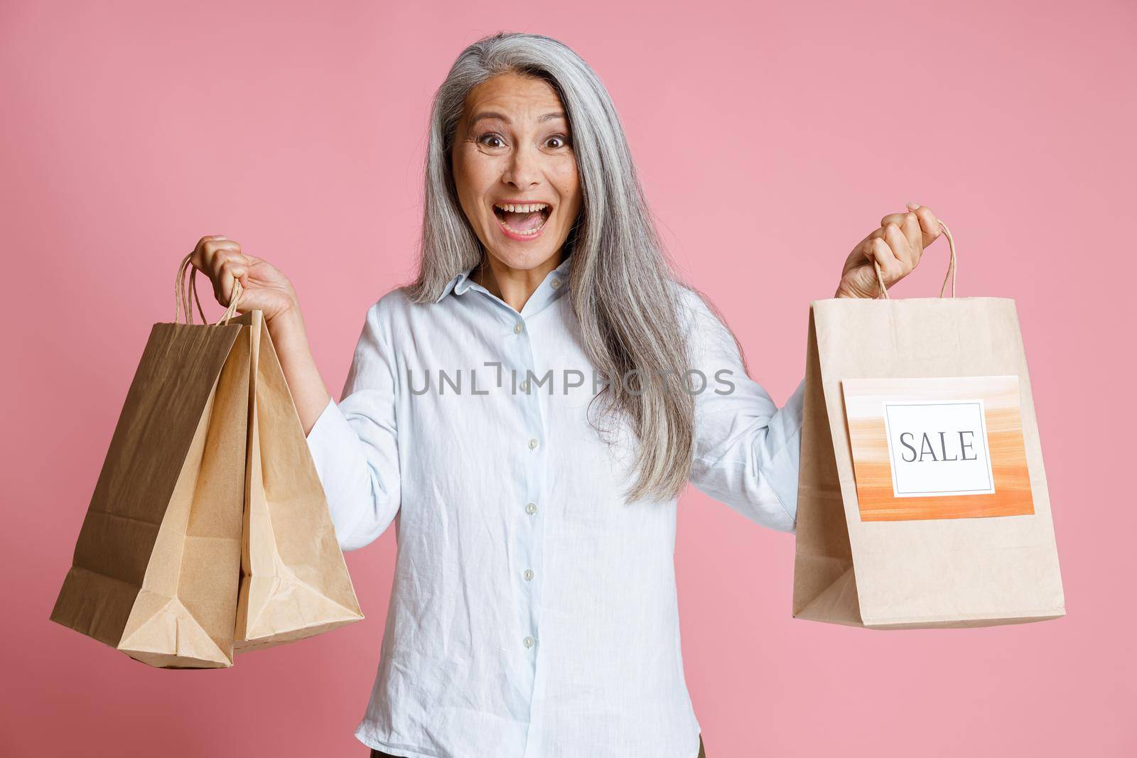 Excited mature Asian woman holds paper shopping bags with colorful Sale sign standing on pink background in studio