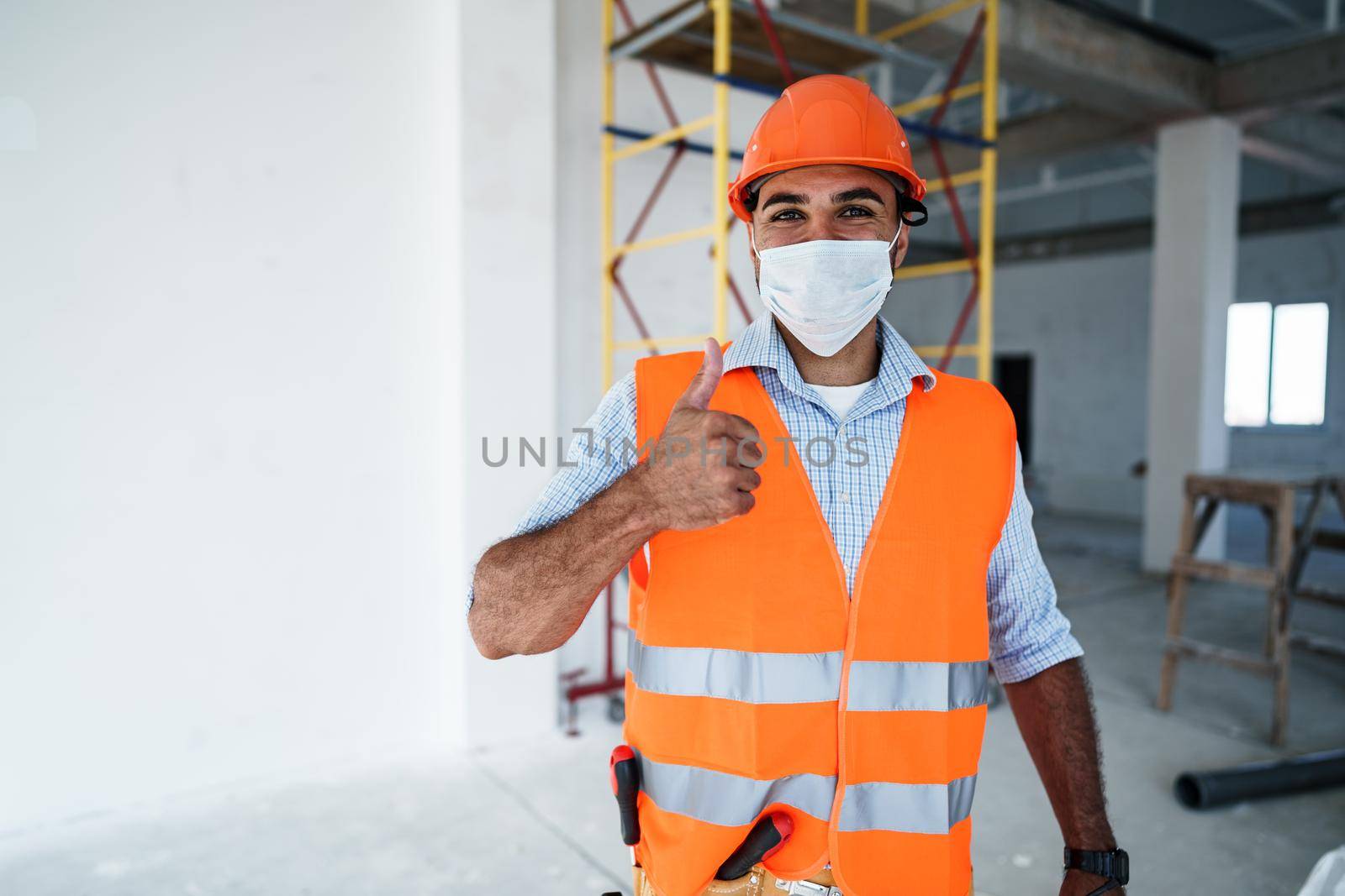 Portrait of mixed race man builder in workwear and hardhat wearing medical mask, close up photo