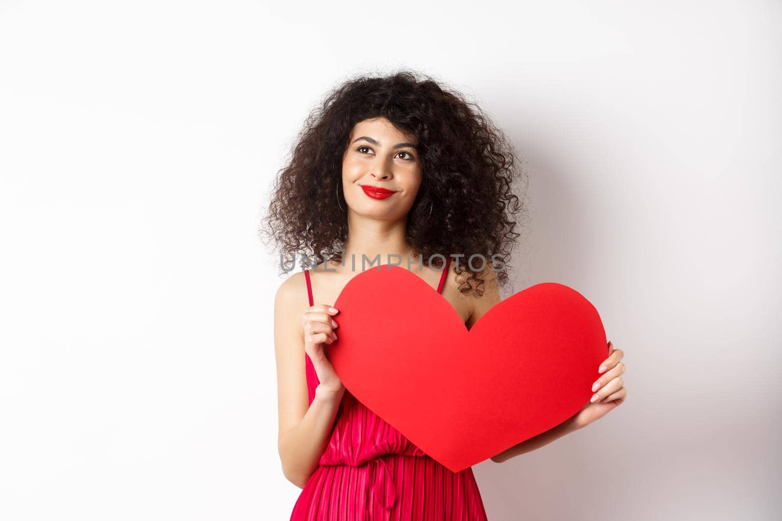 Young romantic woman dreaming about love on Valentines day, looking for soulmate, holding big red heart cutout and gazing pensive left, white background.