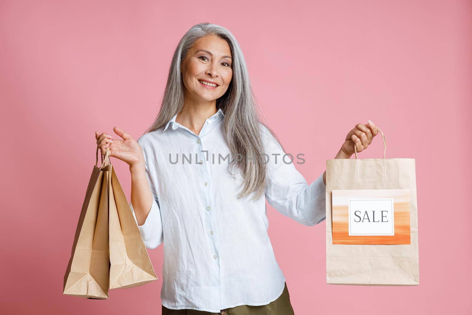 Positive middle aged Asian female model holds paper shopping bags with bright Sale sign standing on pink background in studio