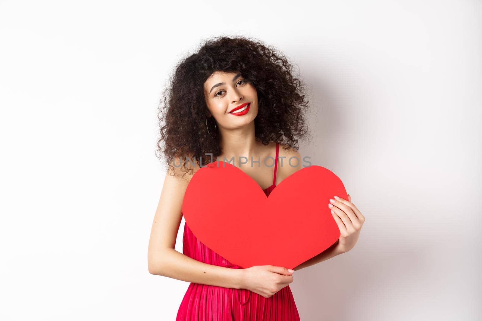 Romantic tender woman with curly hair, hugging big red heart and smiling, look with love, standing against white background.