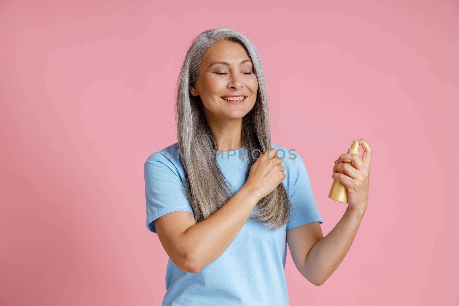 Positive Asian lady applies spray product onto loose grey hair standing on pink background in studio. Mature beauty lifestyle
