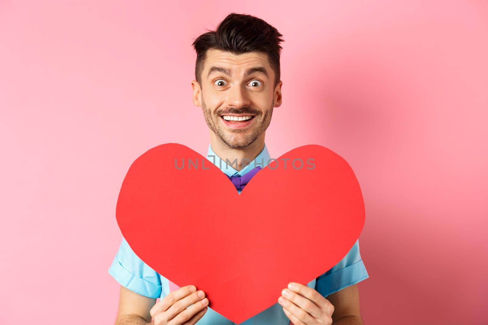 Hopeful man in love showing red heart sign, smiling at camera, waiting for soulmate on Valentines day, standing on pink background by Benzoix