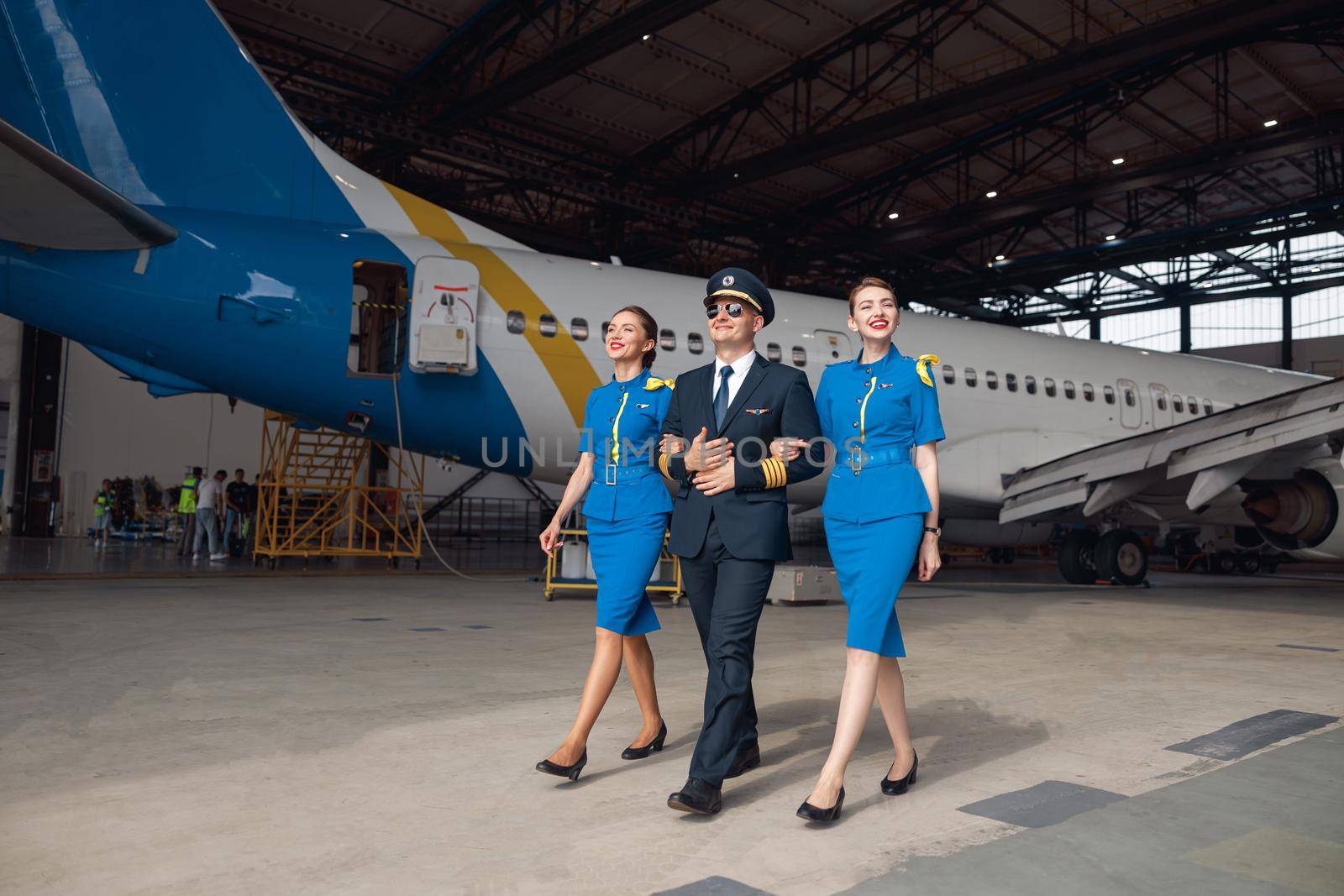 Full length shot of pilot in uniform and aviator sunglasses walking together with two air stewardesses in blue uniform in front of big passenger airplane in airport hangar. Aircraft, occupation