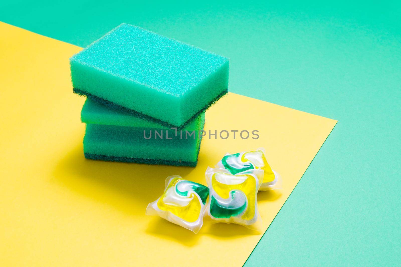 two green sponges for washing dishes and three capsules for dishwasher on a yellow and green background, copy space, blank for design