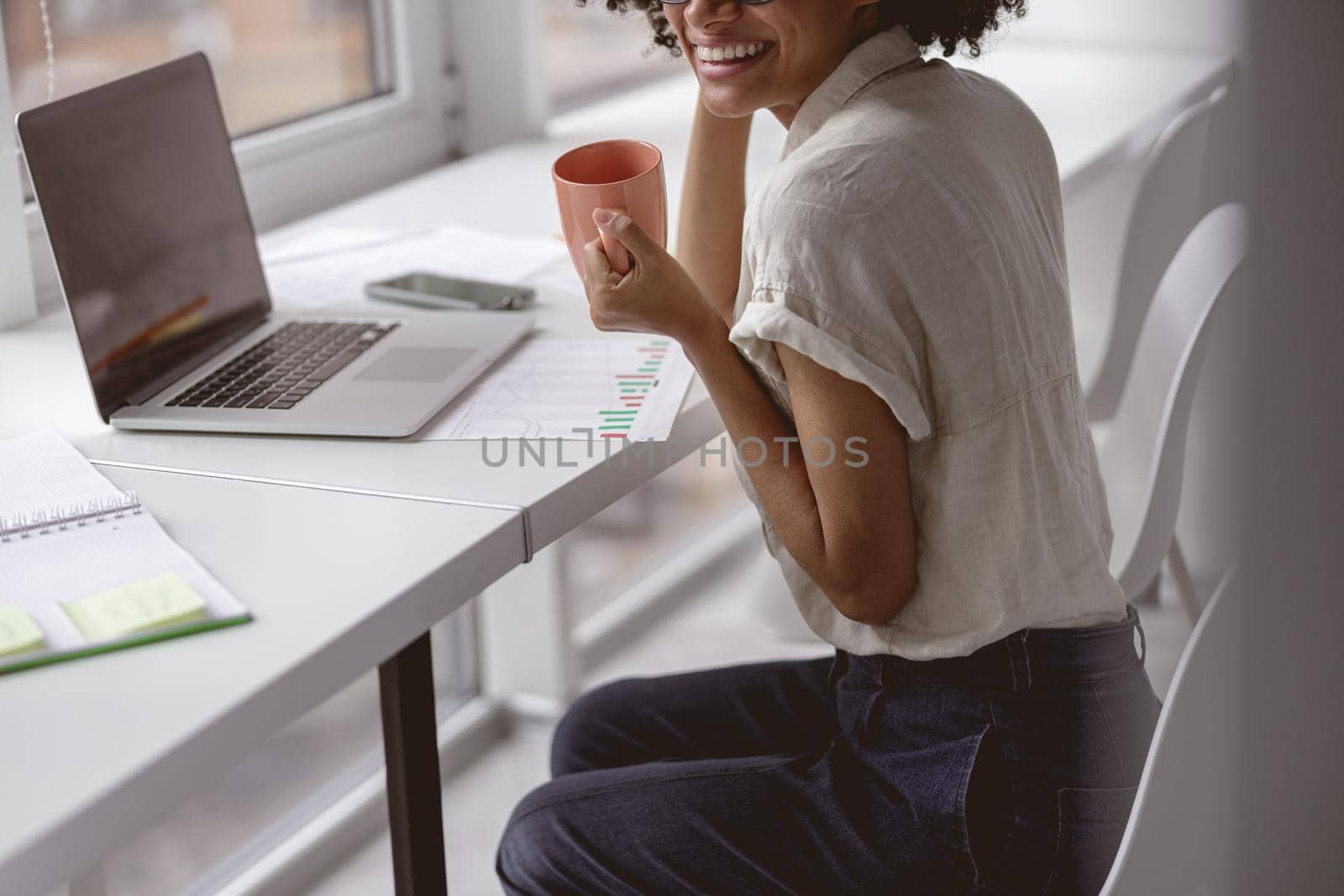 Cropped photot of happy multiethnic female wearing glasses and sitting at the desk with laptop while enjoying coffee
