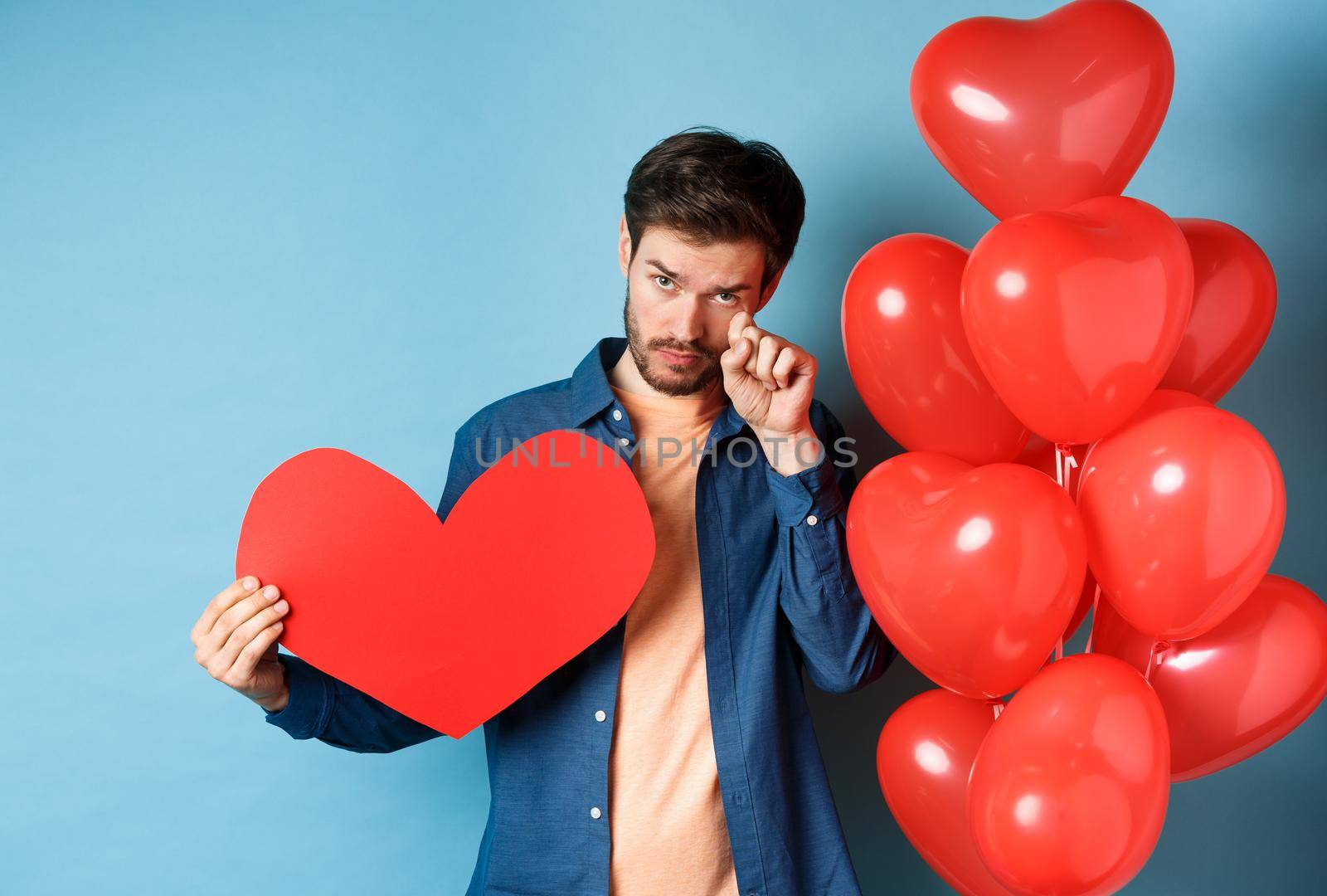 Sad and heartbroken man crying, wiping tears, standing with red heart and balloons, breakup on Valentines day, blue background by Benzoix
