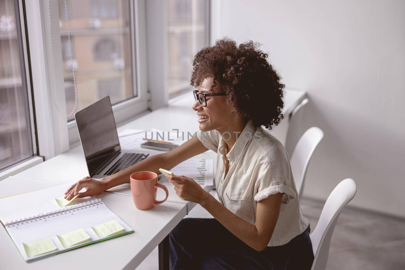 Side view of smiling woman working with laptop and notebook while sitting near the window
