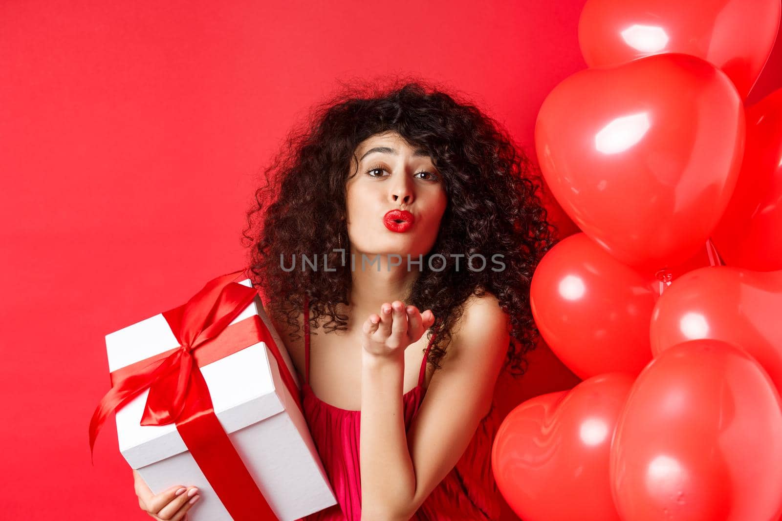 Romance and Valentines day concept. Pretty curly-haired girl in red dress sending her love, blowing air kiss at camera, holding gift from lover, standing near hearts on red background.