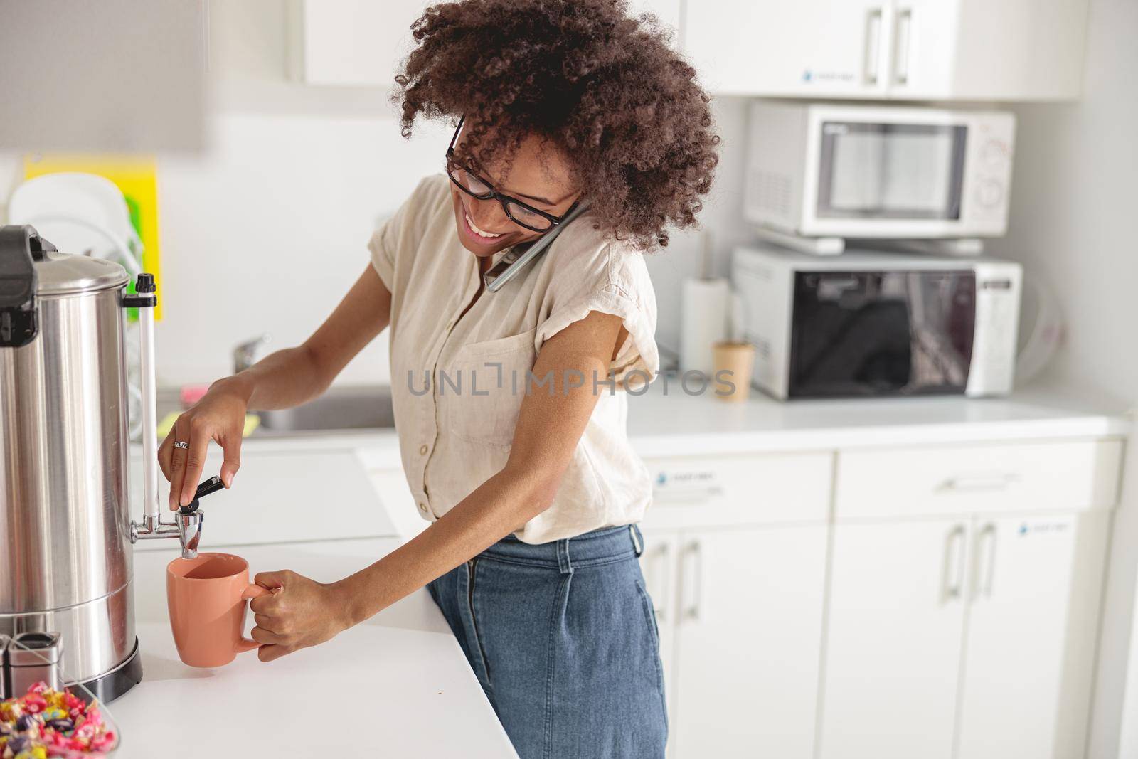 Smiling Afro American woman preparing hot drink while talking on smartphone on office kitchen