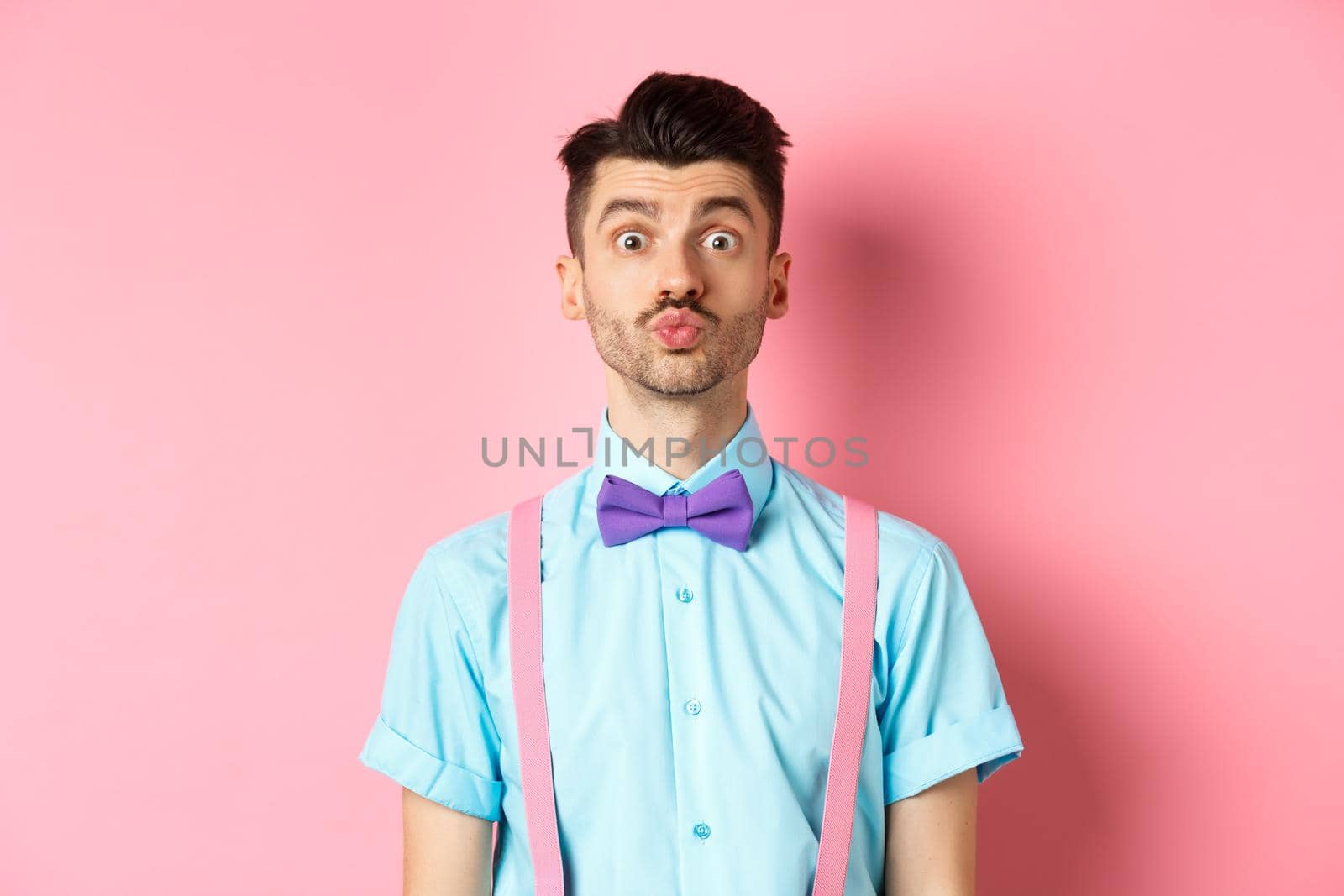 Funny young man waiting for kiss with puckered lips and silly face, standing on romantic pink background. Concept of love and Valentines day.