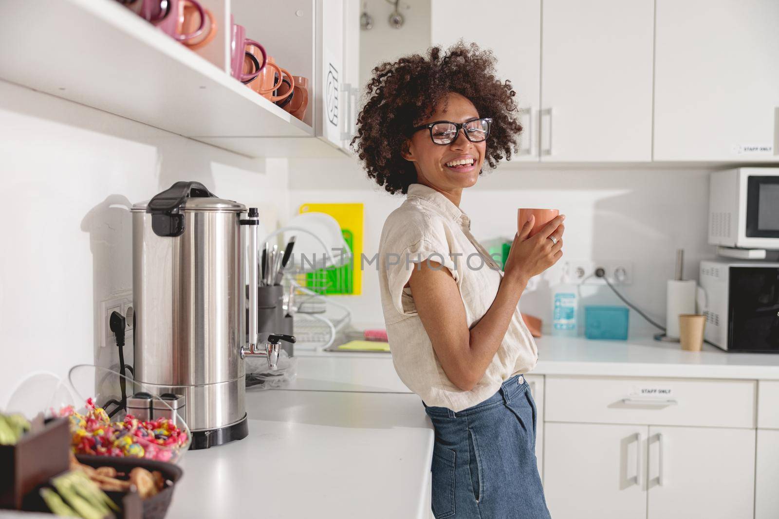 Pretty young woman holding coffee and posing on office kitchen while looking at camera