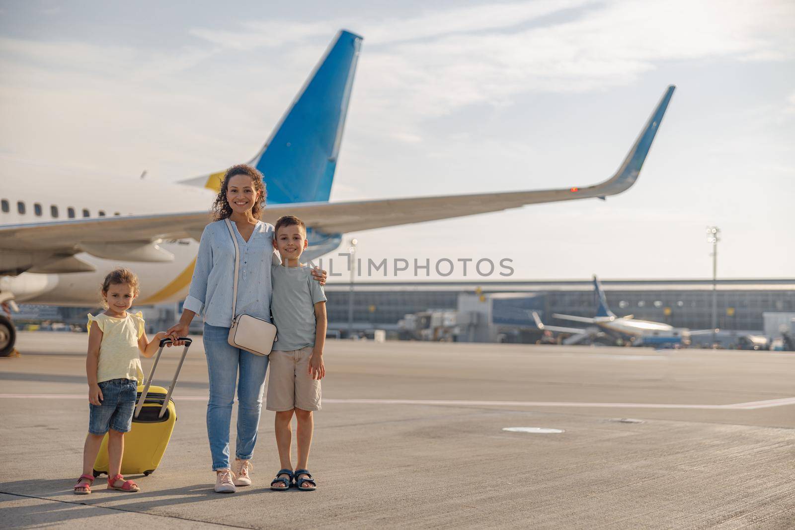 Full length shot of happy mother with two little kids smiling at camera while standing in front of big airplane in the daytime. Family, vacation, traveling concept