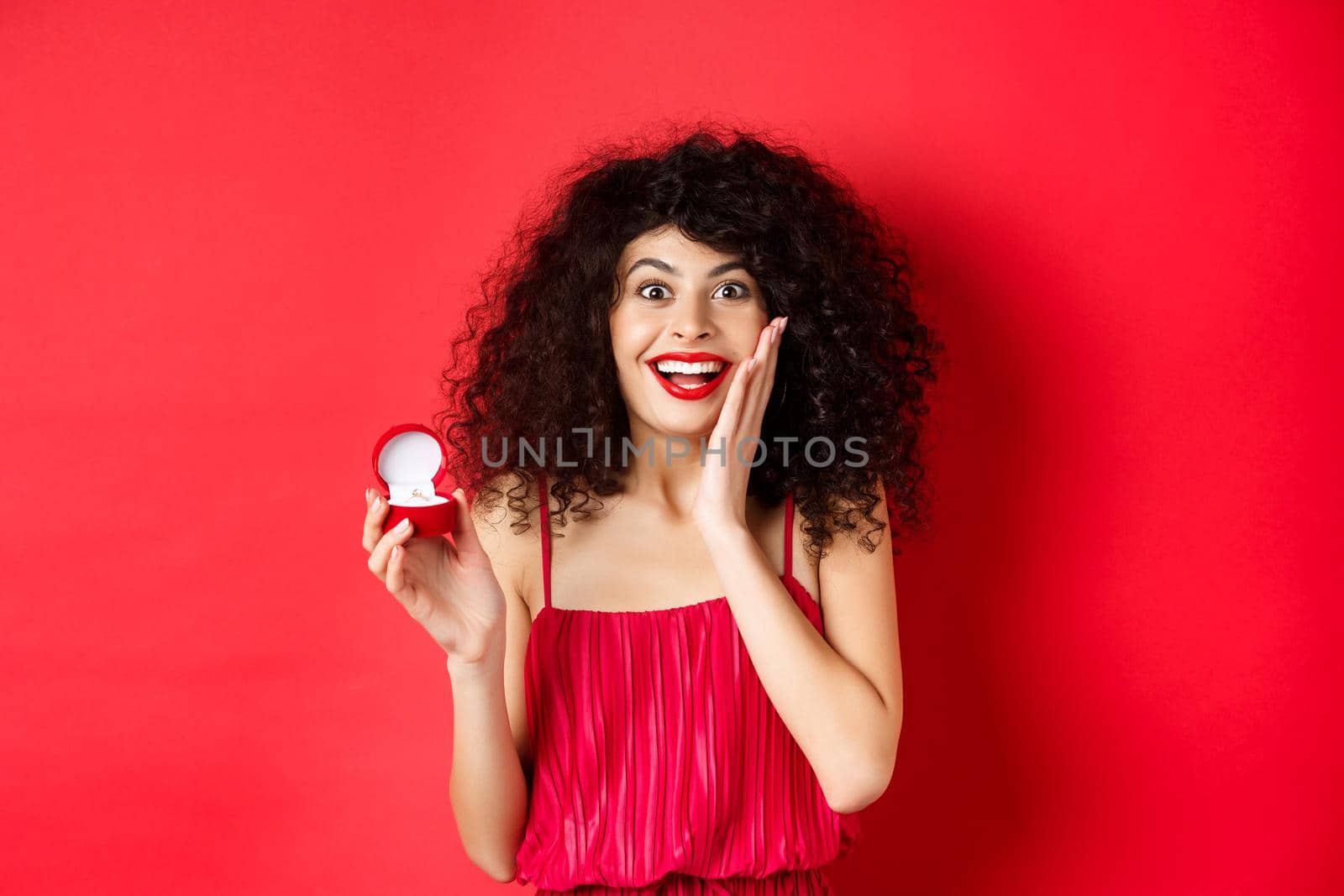 Image of surprised young woman with curly hairstyle, wearing red dress and lipstick, showing engagement ring, going to get married, standing on studio background.