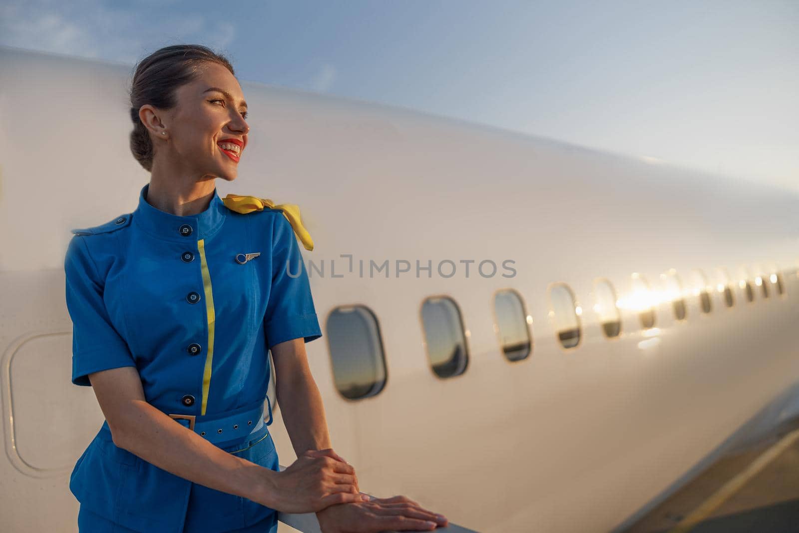 Inspired air hostess in blue uniform smiling aside, standing outdoors at the sunset. Commercial airplane in the background. Aircrew, occupation concept