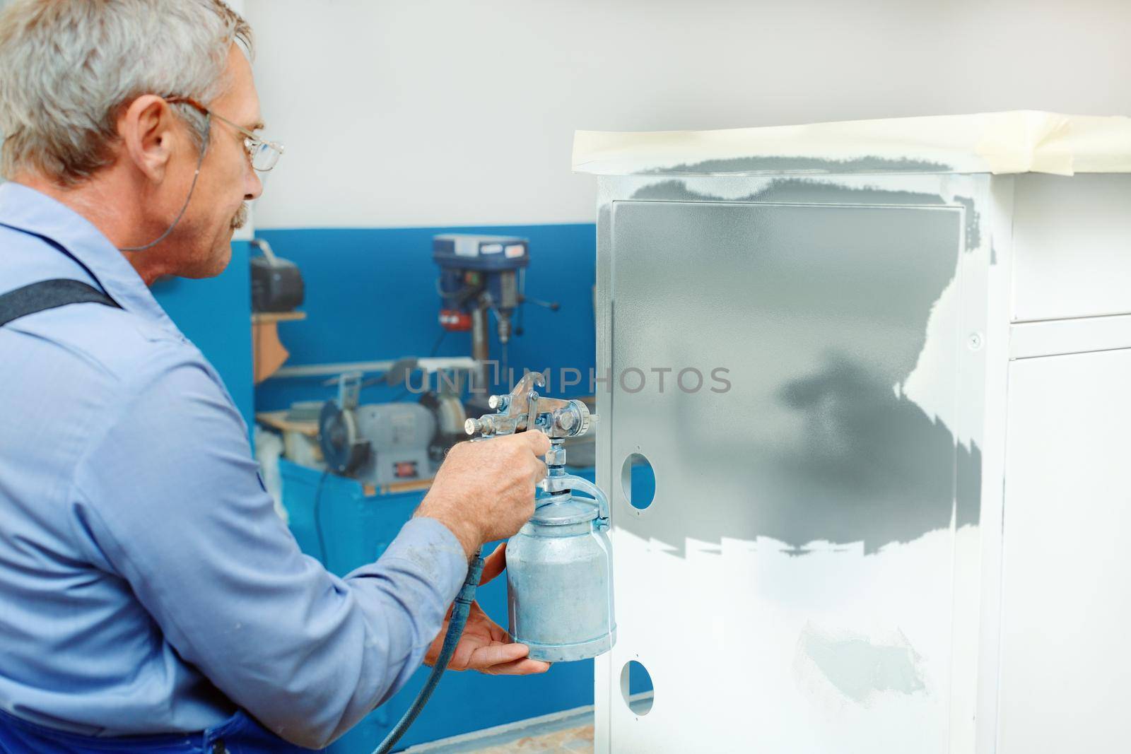 An employee paints metal products. An elderly man with glasses holds a compressor spray gun in his hand and paints a Cabinet in the workshop