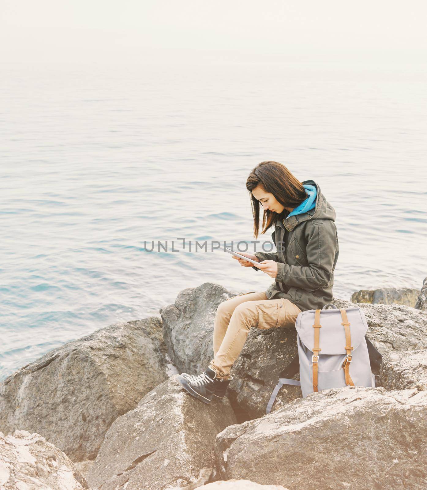 Freelancer and traveler young woman sitting on stone coast near the sea and working on digital tablet.