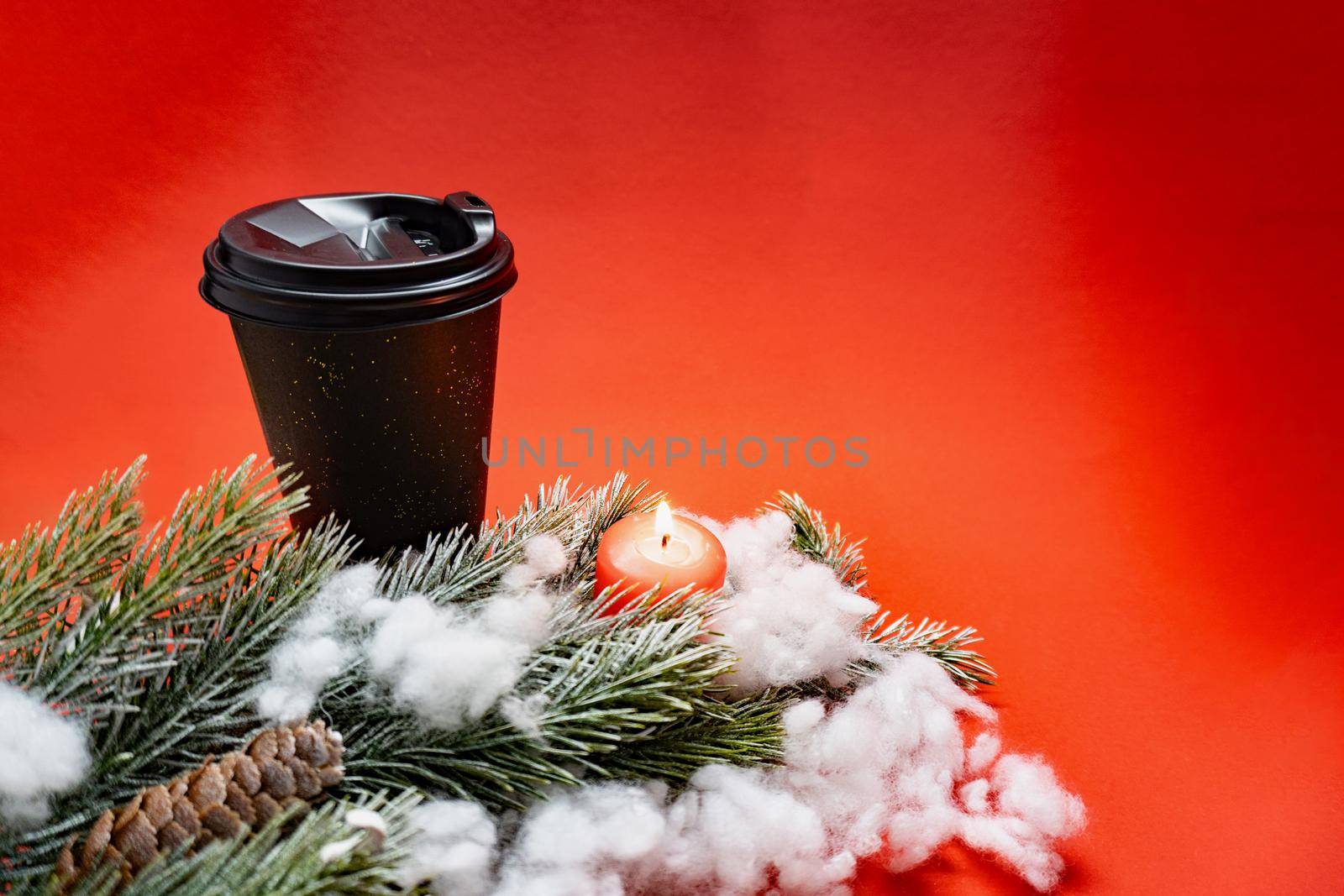 Black coffee cup with plastic cap, green braches with knar and snow and a red candle on red background with lights, new year concept, place for the text.