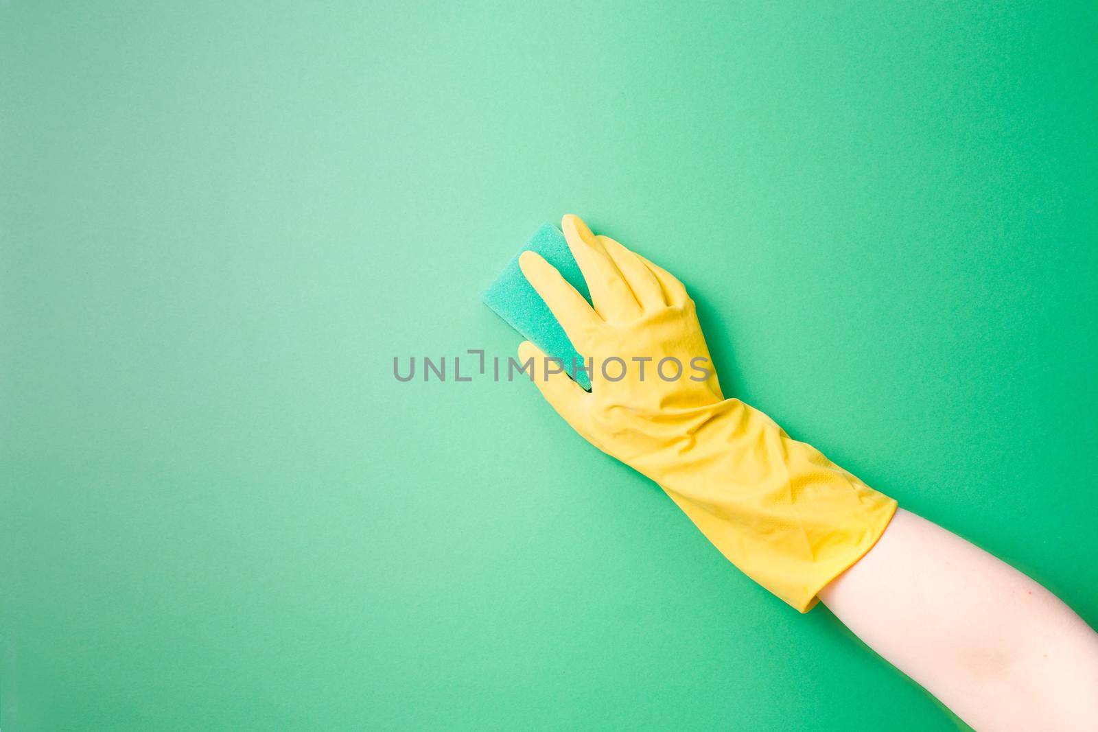 a female hand in a yellow rubber glove washes a plain surface with a green paralon sponge for washing dishes and cleaning, green background copy space, cleaning company and spring cleaning concept