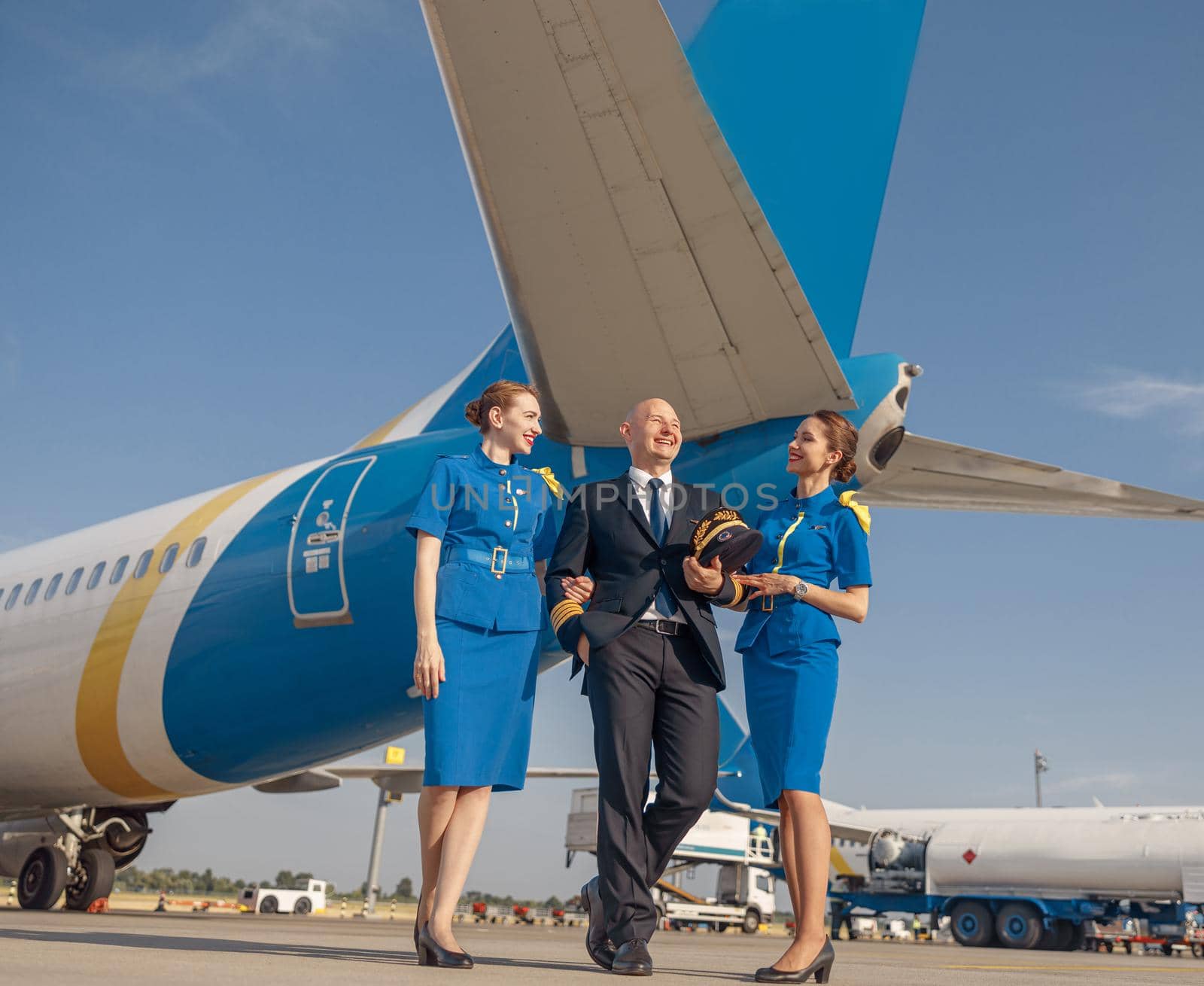 Full length shot of happy pilot and two pretty stewardesses standing together in front of an airplane and smiling after landing. Aircraft, aircrew, occupation concept