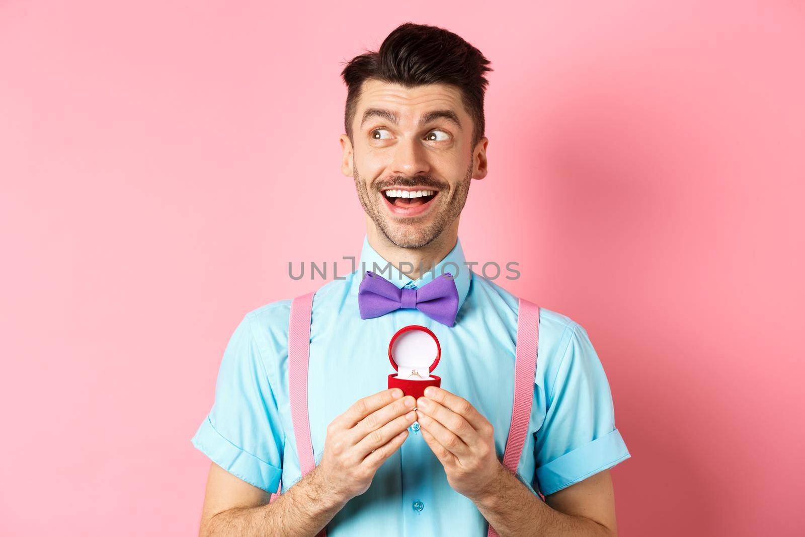 Valentines day. Romantic handsome man looking dreamy and smiling, showing engagement ring for his lover, standing happy over pink background.