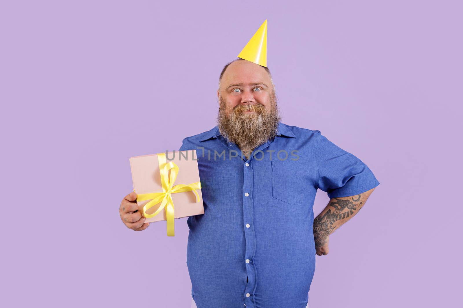 Mature man with overweight with party hat holds gift box on purple background by Yaroslav_astakhov