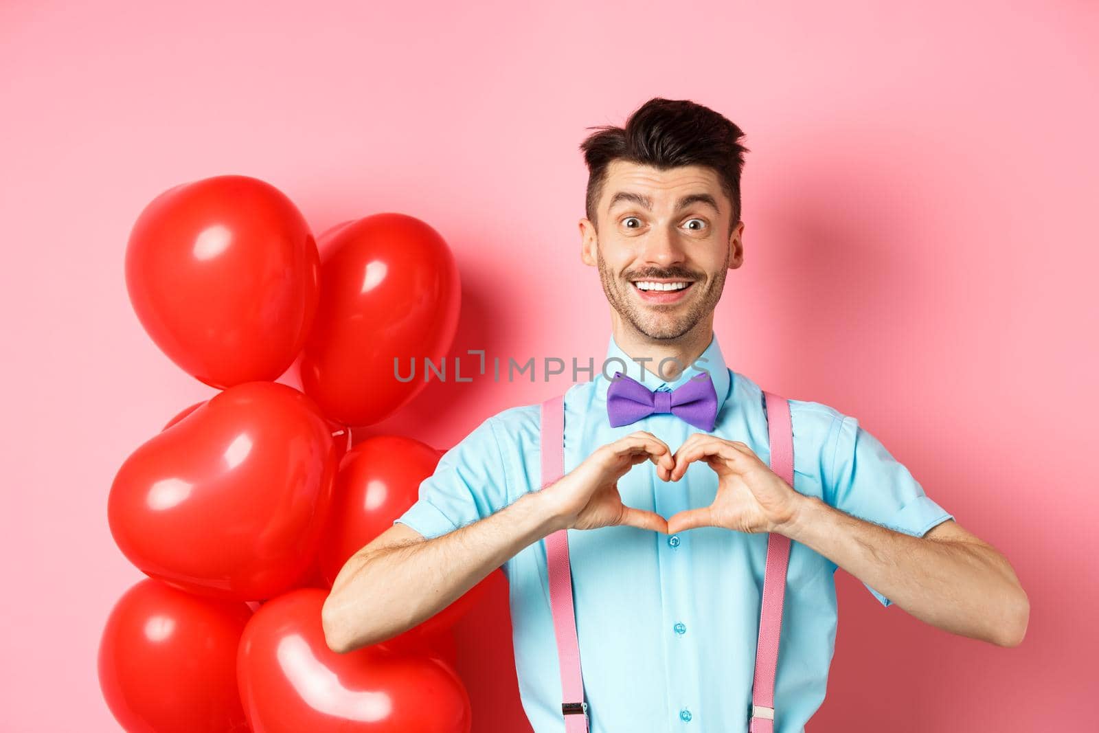 Valentines day concept. Romantic guy in bow-tie, showing heart gesture and say I love you with hopeful smile, standing on pink background near balloons by Benzoix