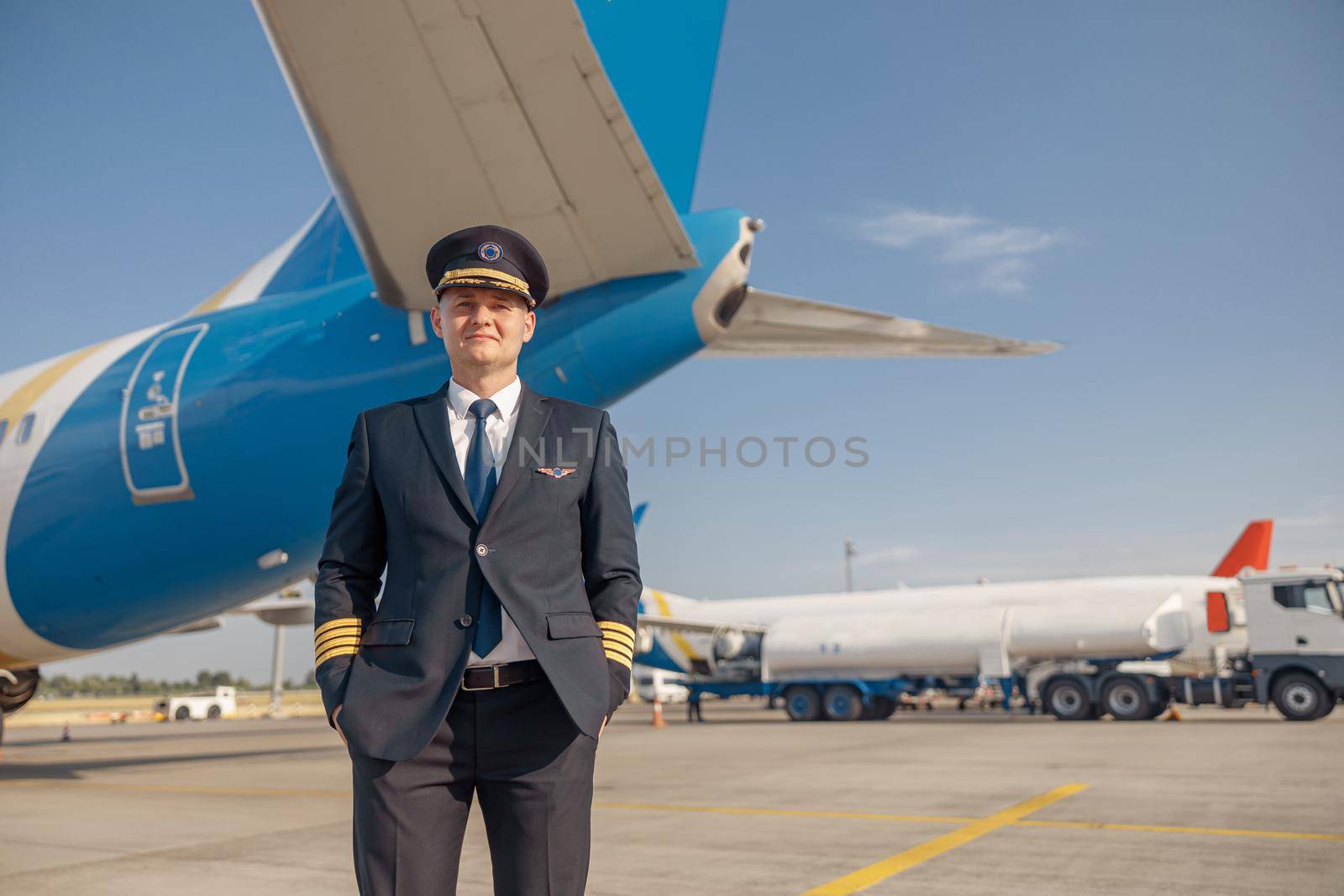 Professional pilot in uniform looking at camera, standing in front of big passenger airplane ready for departure in airport. Aircraft, occupation, transportation concept