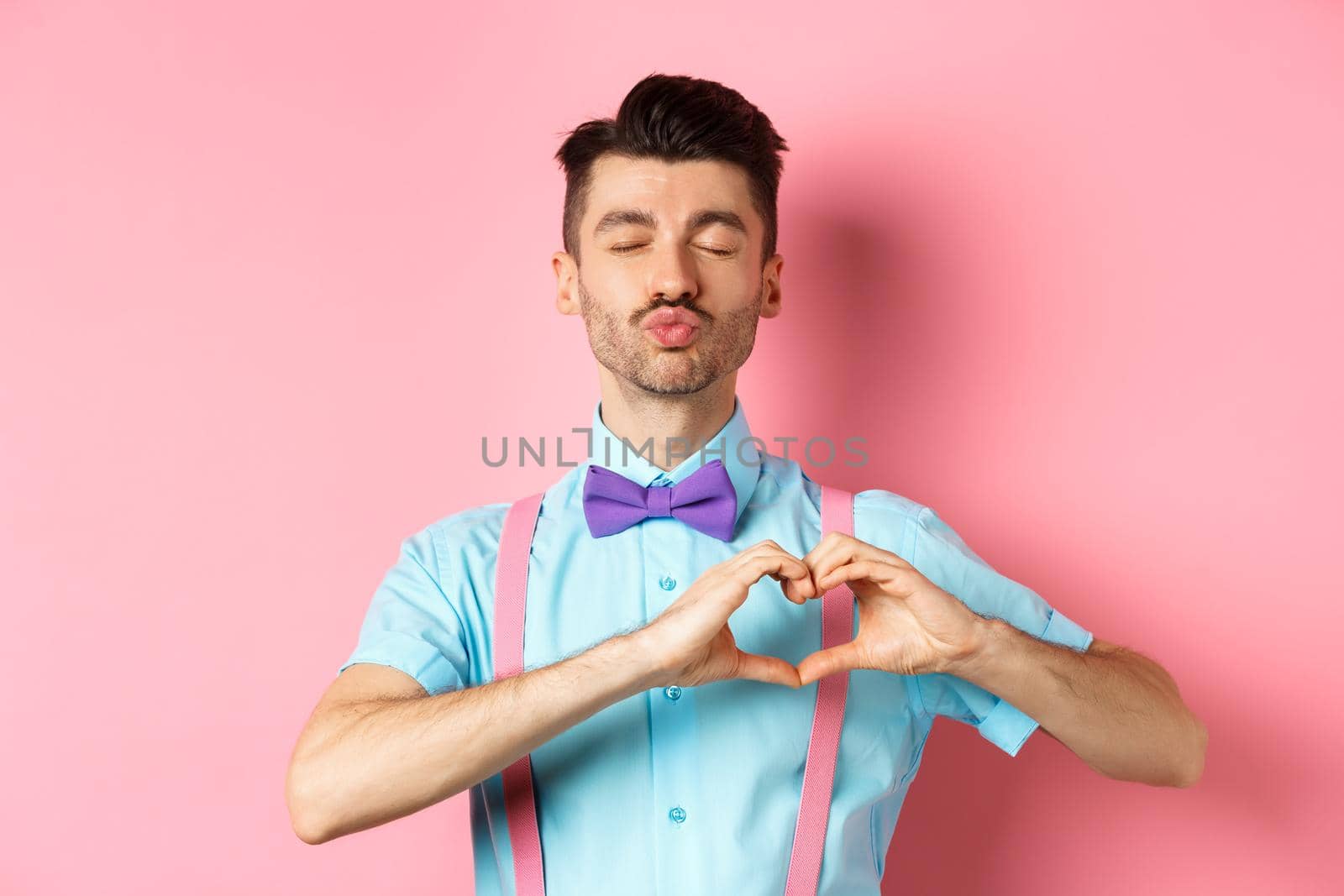 Romantic man in funny bow-tie waiting for kiss, close eyes and pucker lips kissing, showing heart gesture, being in love on Valentines day, standing over pink background by Benzoix