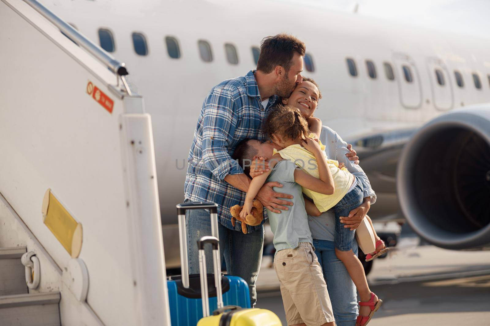 Family of four kissing each other while going on a trip, standing in front of big airplane outdoors. People, traveling, vacation concept