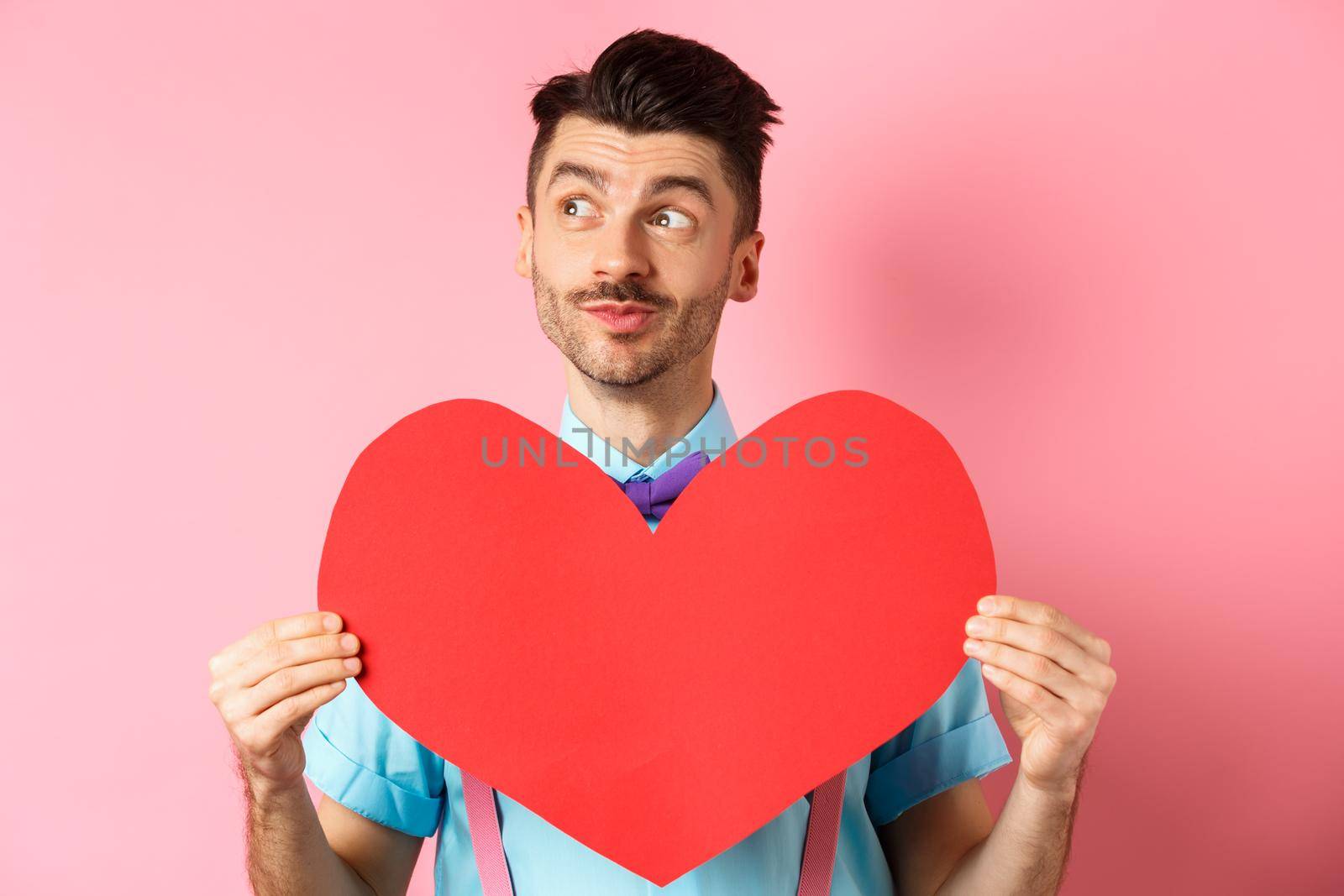 Valentines day concept. Dreamy romantic man looking left and showing big red heart cutout, standing on pink background by Benzoix