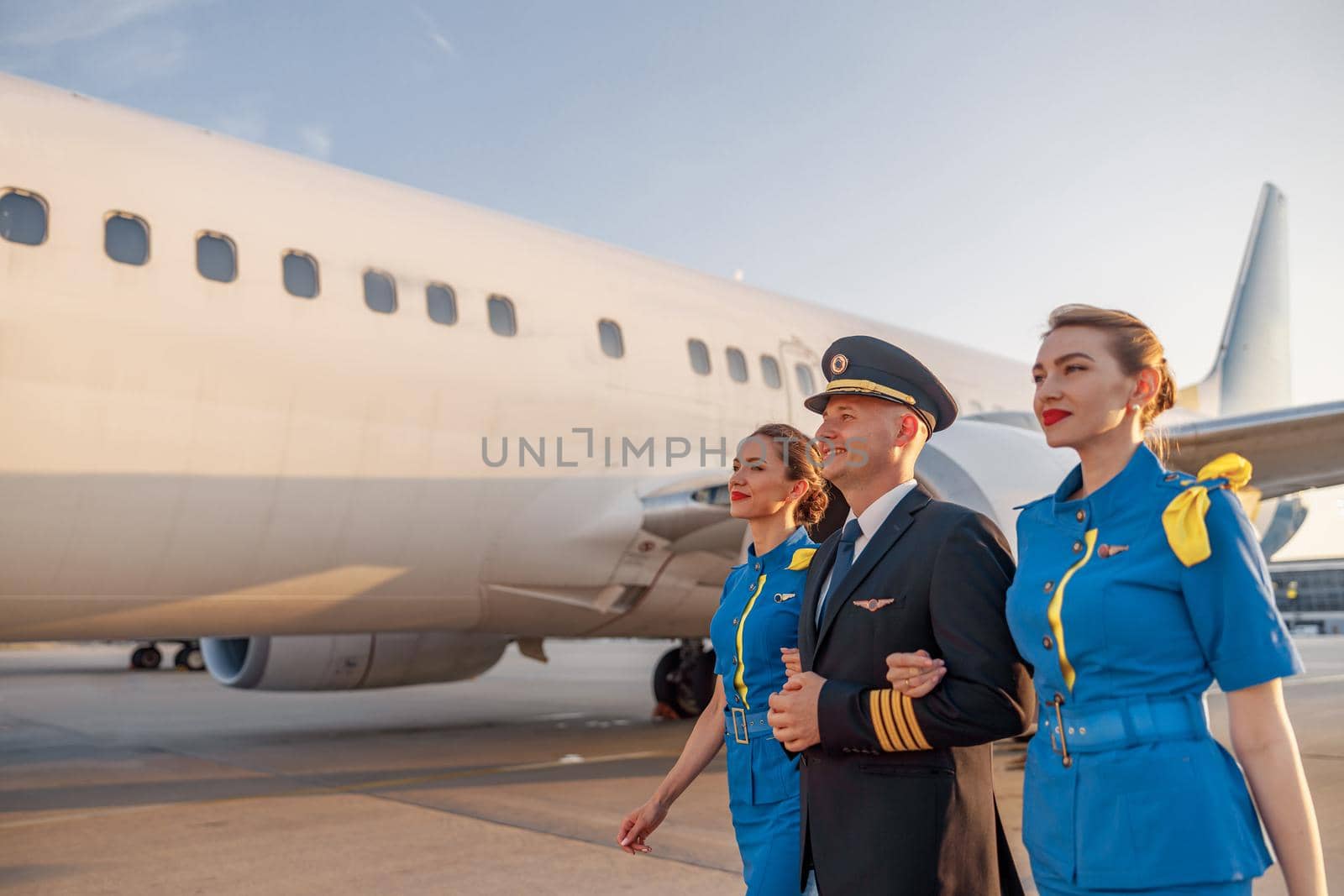 Excited male pilot walking together with two flight attendants in blue uniform in front of an airplane in terminal at sunset by Yaroslav_astakhov