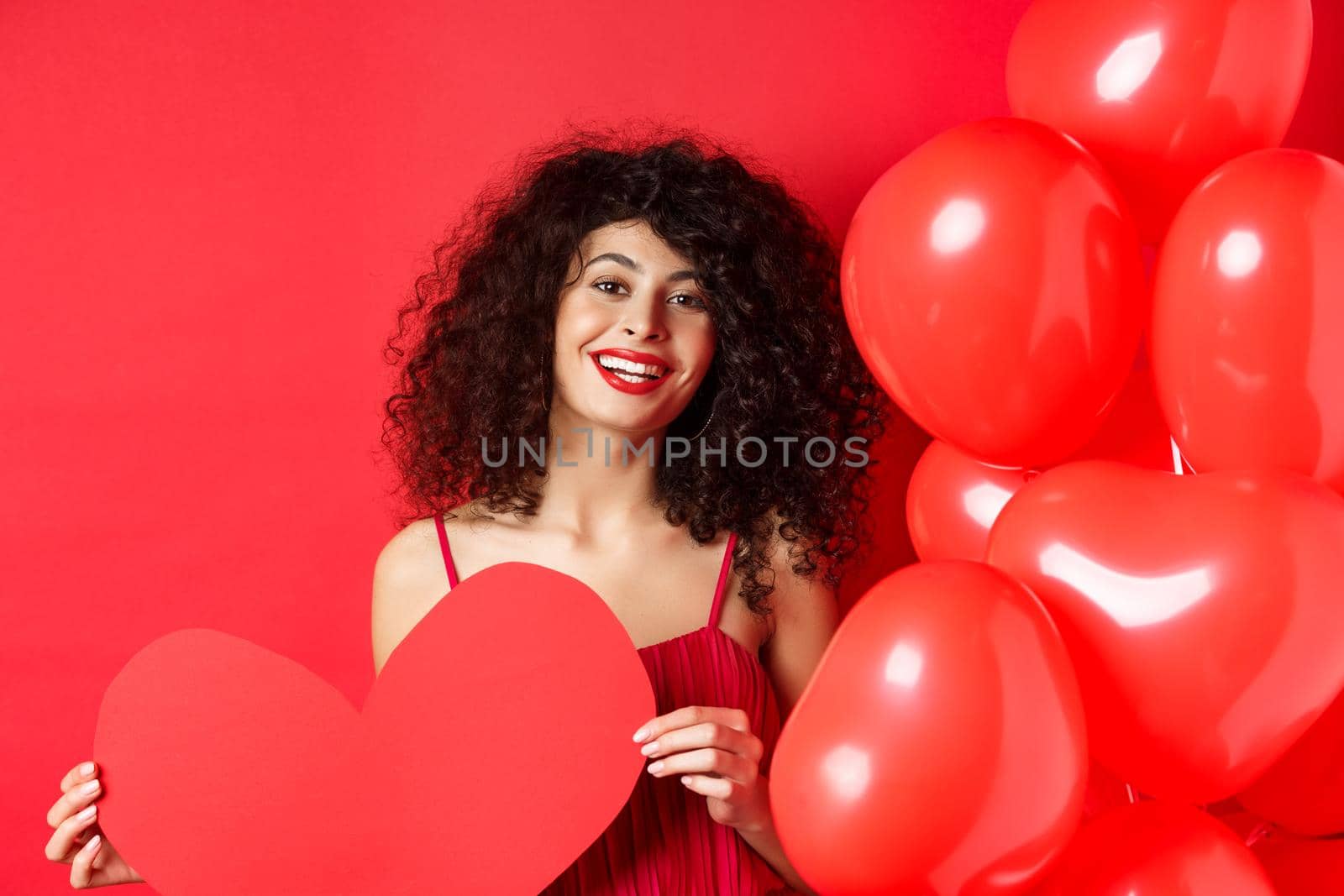 Happy valentines day. Romantic girl in dress waiting for true love, showing big red heart and standing near balloons on red background.
