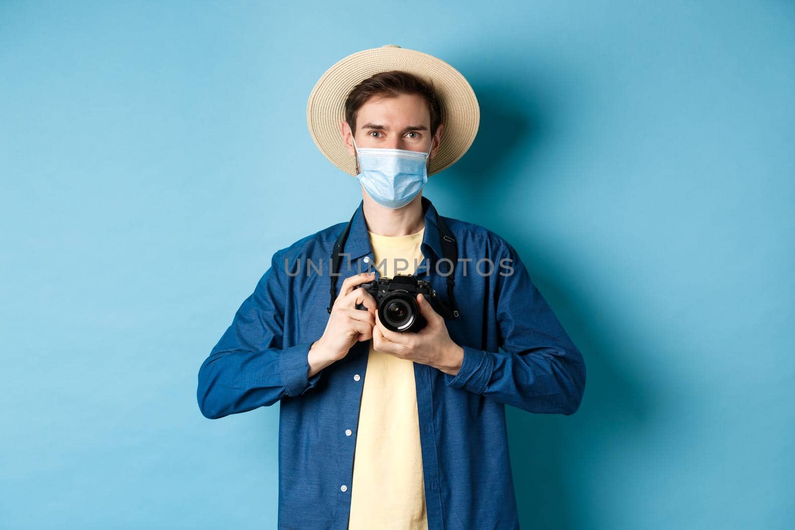 Covid-19, pandemic and travel concept. Happy guy on vacation taking pictures with camera, wearing medical mask and summer hat, blue background.