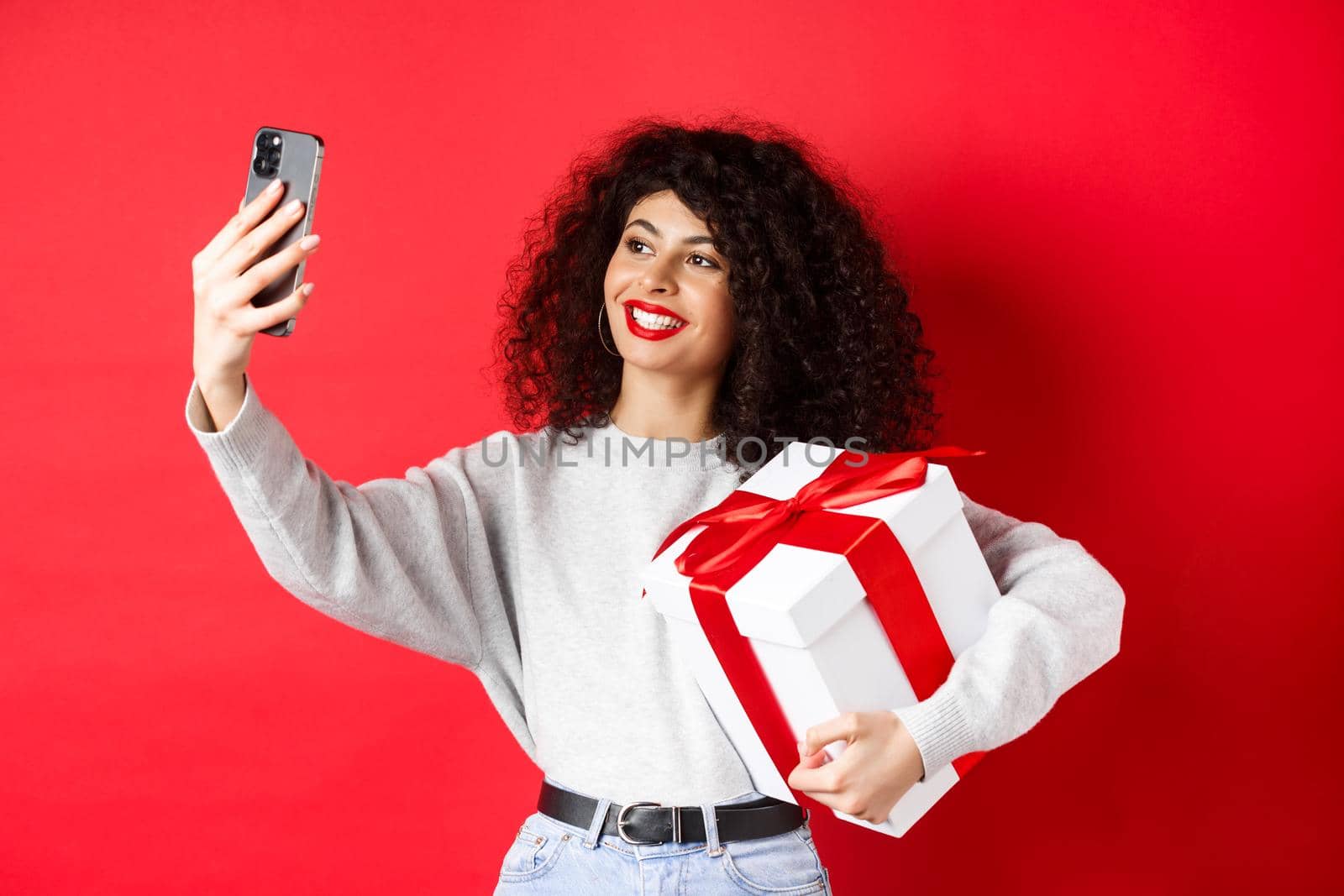 Happy young woman taking selfie with her valentines day gift, holding present and photographing on smartphone, posing on red background by Benzoix