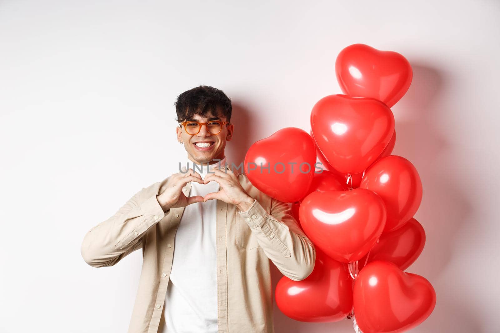 Valentines day and romance concept. Handsome young man standing near red hearts balloons and showing heart gesture, being in love and smiling, white background by Benzoix
