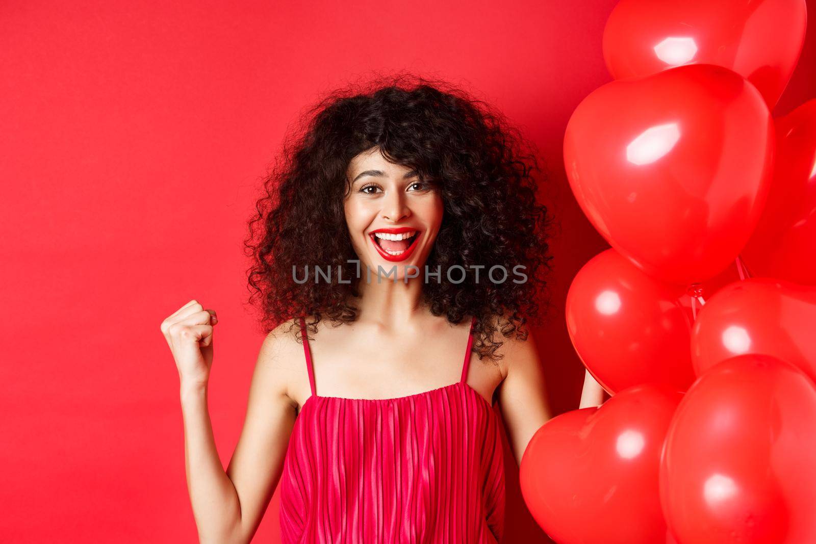 Excited pretty woman winning, say yes and smiling, celebrating success, making fist pump motivated, standing near heart balloons and red background.