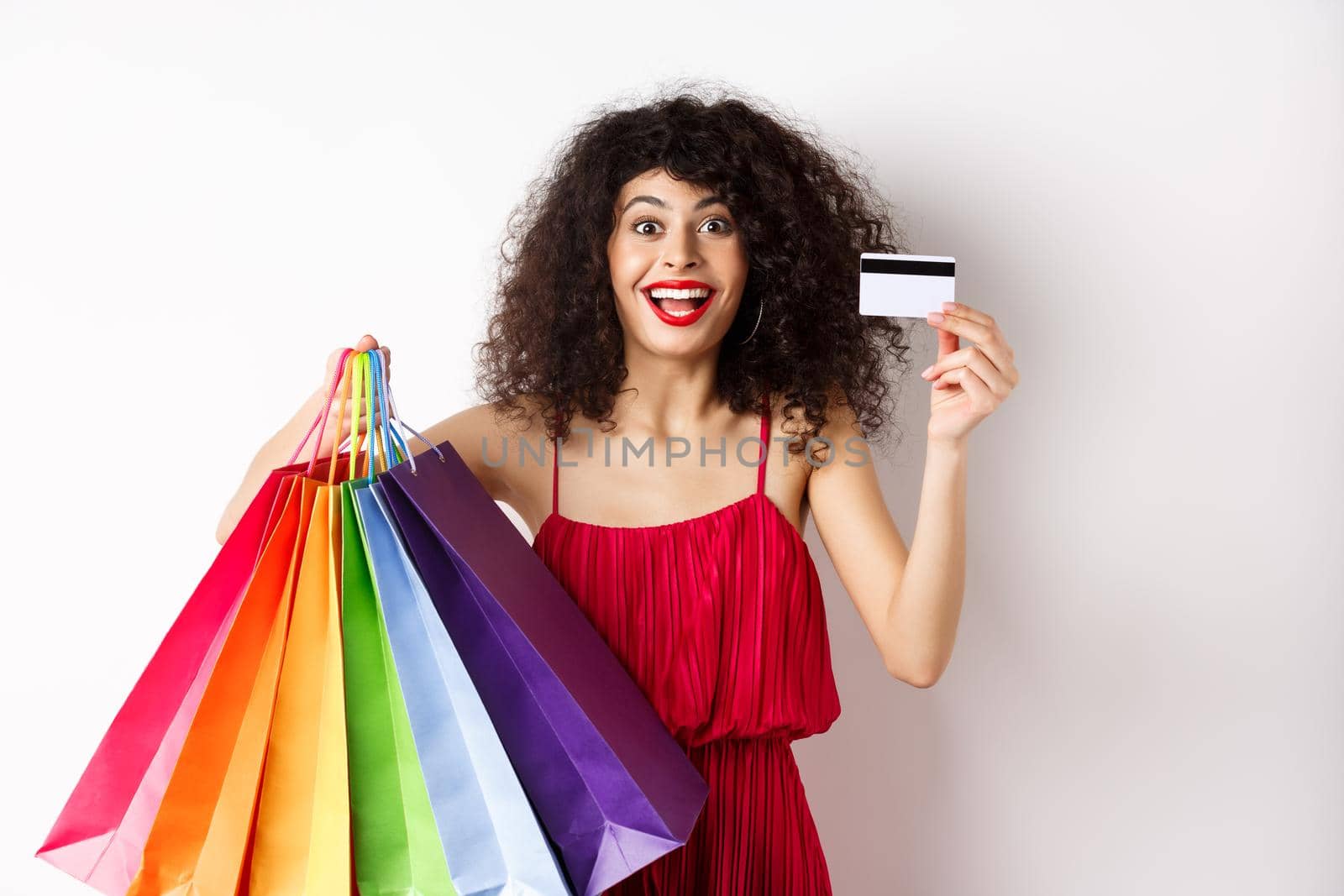 Stylish caucasian woman with curly hair and red dress, showing shopping bags and her plastic credit card, smiling amused, standing over white background.