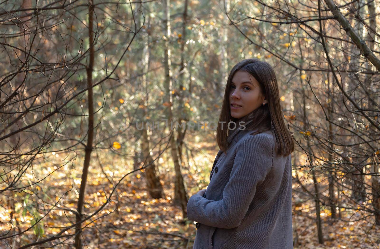 Half-turned girl in a gray coat, which stands in the forest on a sunny autumn day