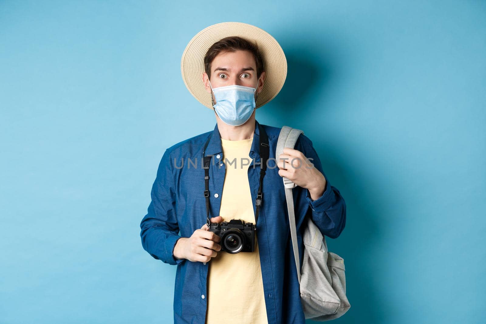 Covid-19 and summer holidays concept. Excited tourist in straw hat and medical mask looking in awe, holding camera, taking pictures on holiday during pandemic, blue background by Benzoix