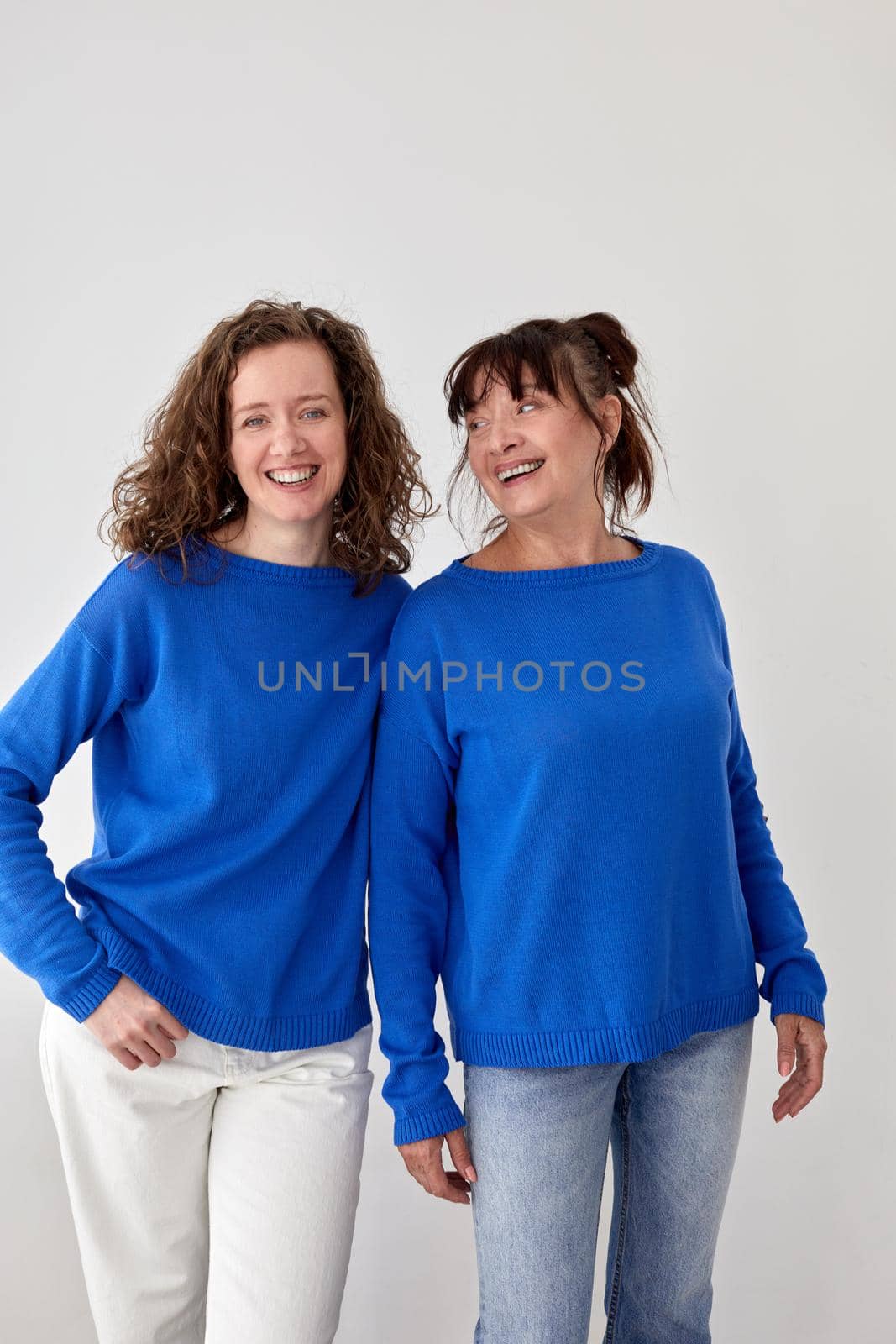 Glad loving mature mother and adult daughter wearing similar blue sweaters on gray background in studio