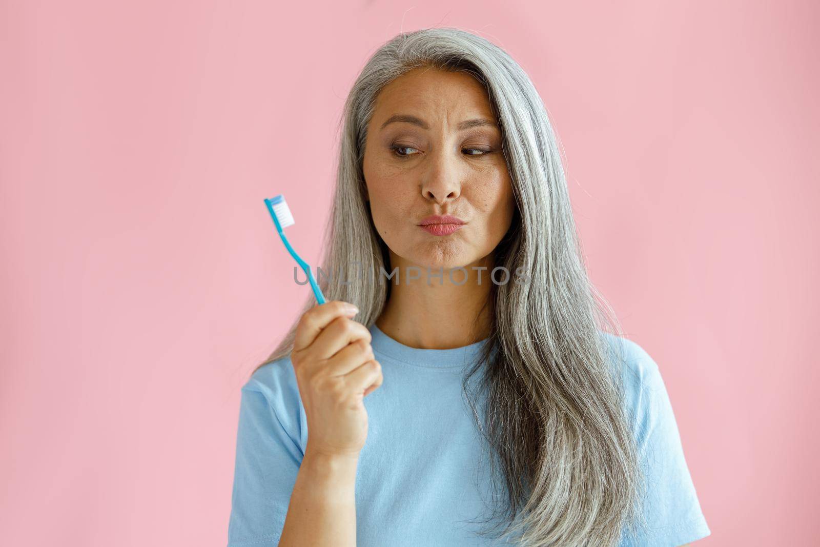 Doubting silver haired mature Asian lady in blue t-shirt looks at toothbrush on pink background in studio. Oral cavity hygiene