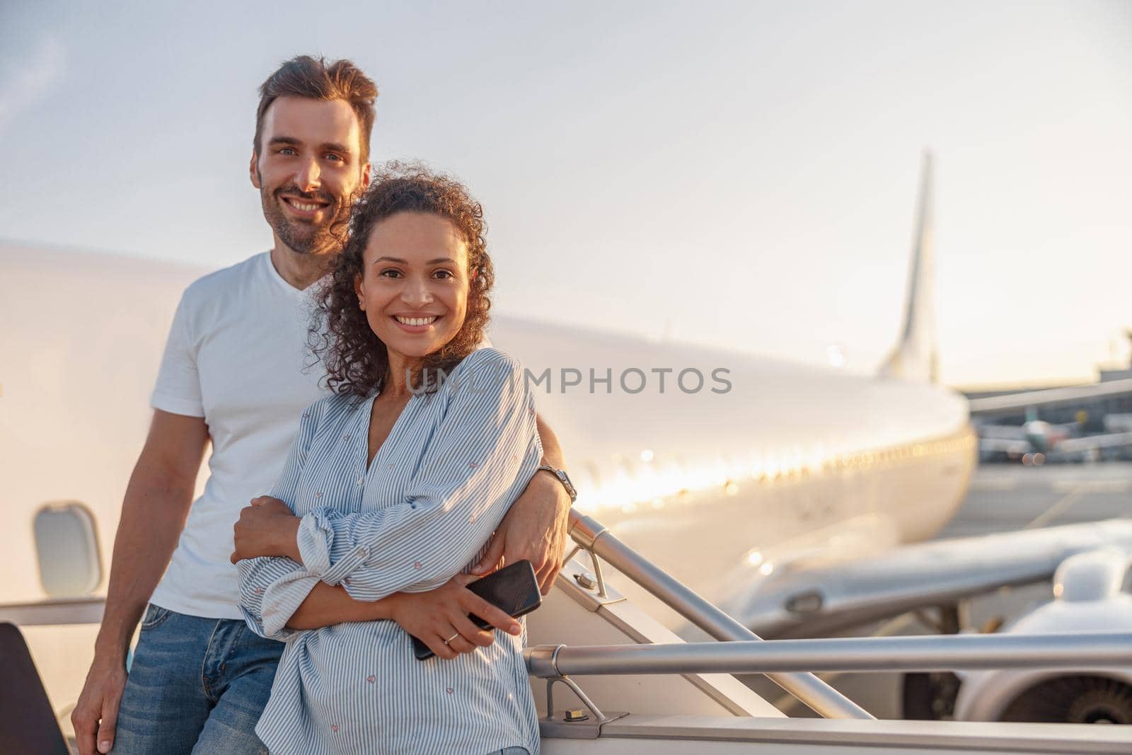Portrait of happy couple of tourists, man and woman looking excited while standing together outdoors ready for boarding the plane at sunset by Yaroslav_astakhov