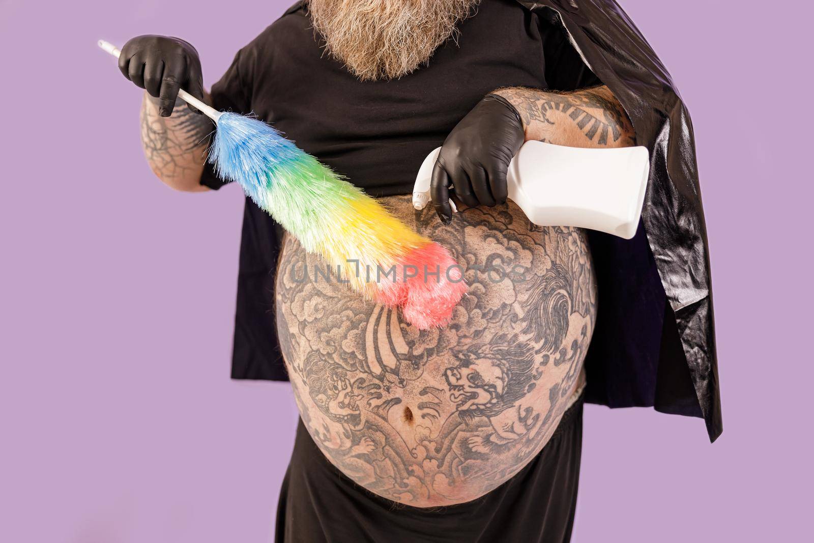 Fat man in hero suit with large bare tattooed tummy sprays detergent onto colorful brush from bottle on purple background in studio closeup