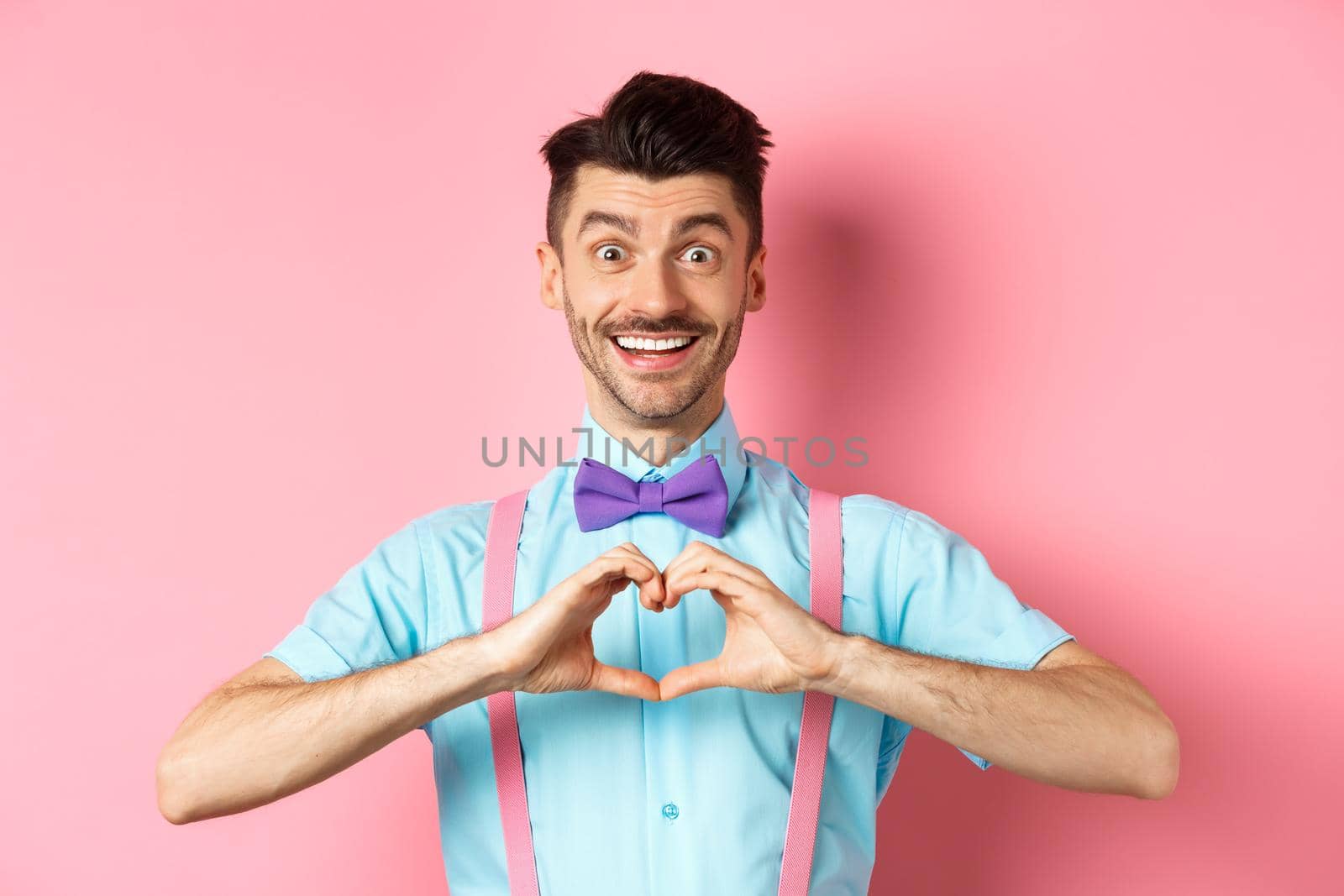 Happy Valentines day. Smiling funny guy with moustache showing heart sign and looking at lover, saying I love you, express feeling, standing over romantic pink background.