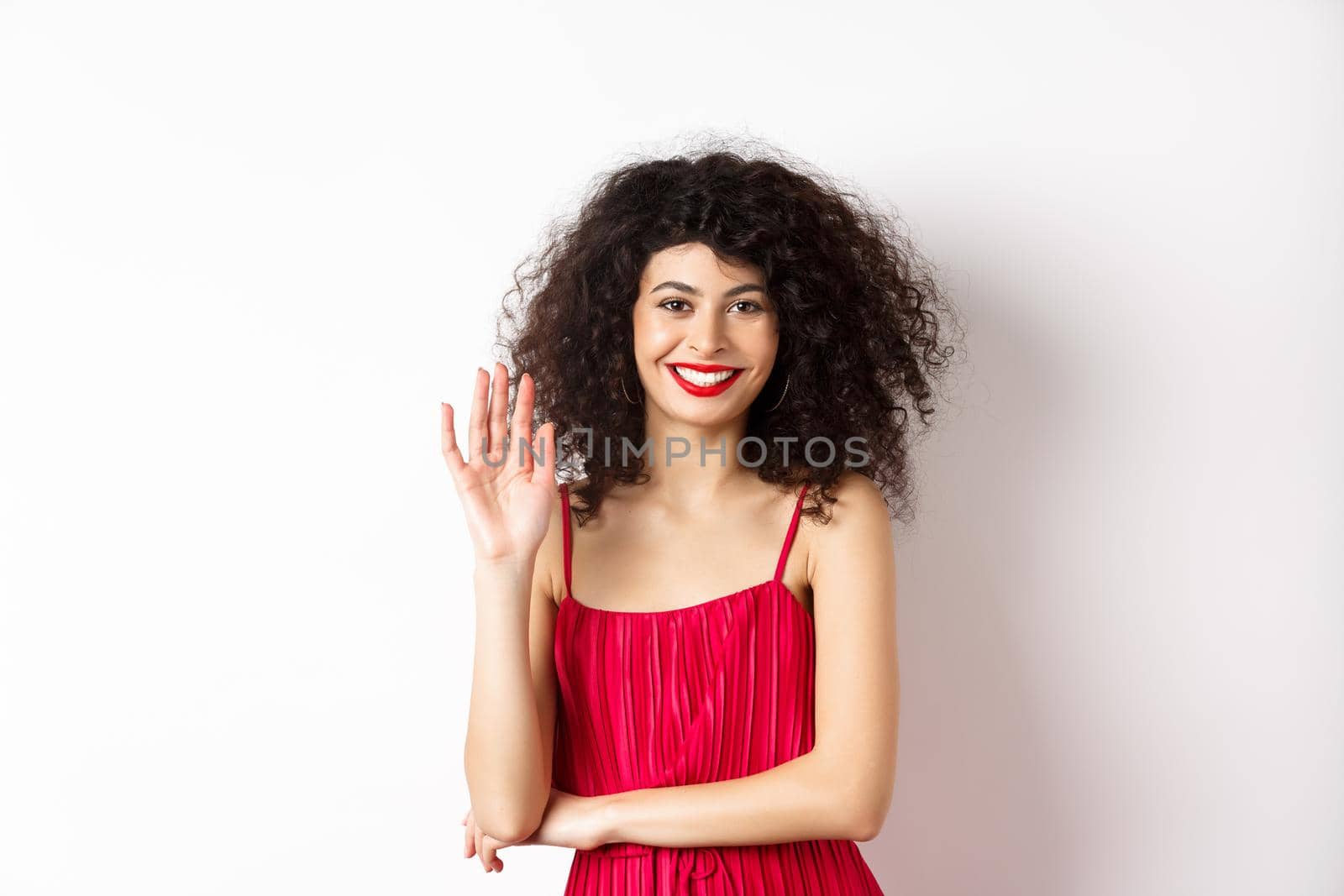Cheerful elegant woman saying hello, waving hand and smiling at camera, greeting someone, standing in red dress on white background by Benzoix