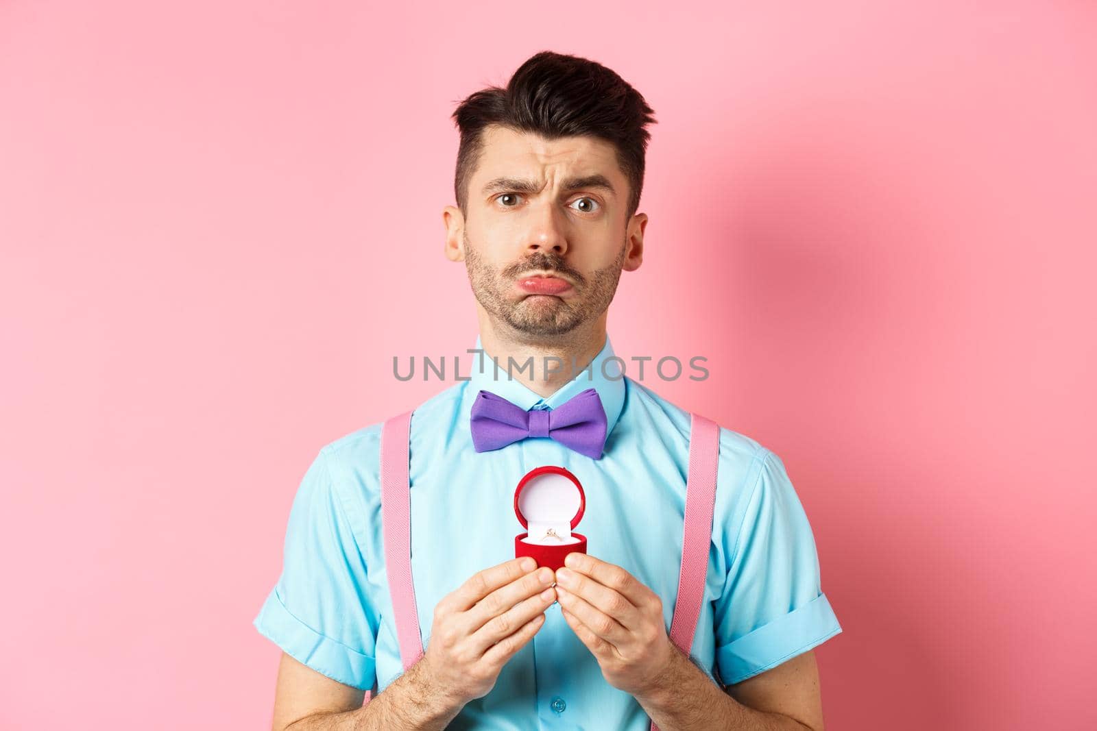 Valentines day. Sad boyfriend being rejected, showing engagement ring and sulking upset, she said no, standing over pink background.