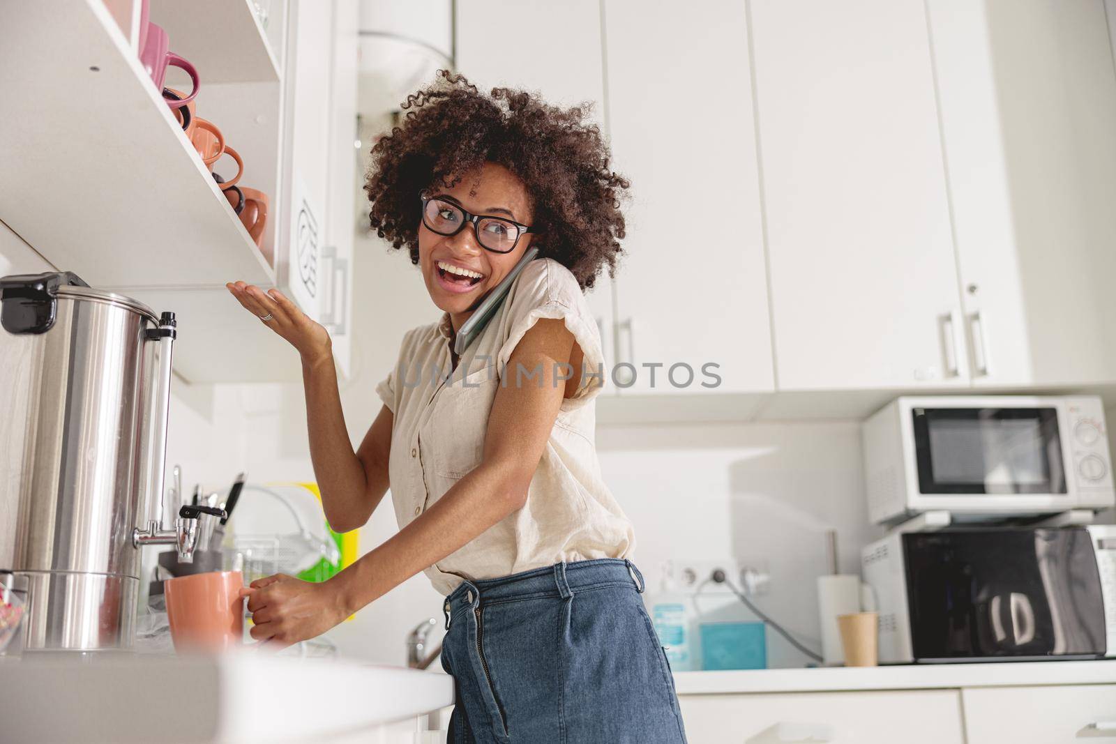 Happy pretty woman making coffee and having a conversation by mobile phone on office kitchen